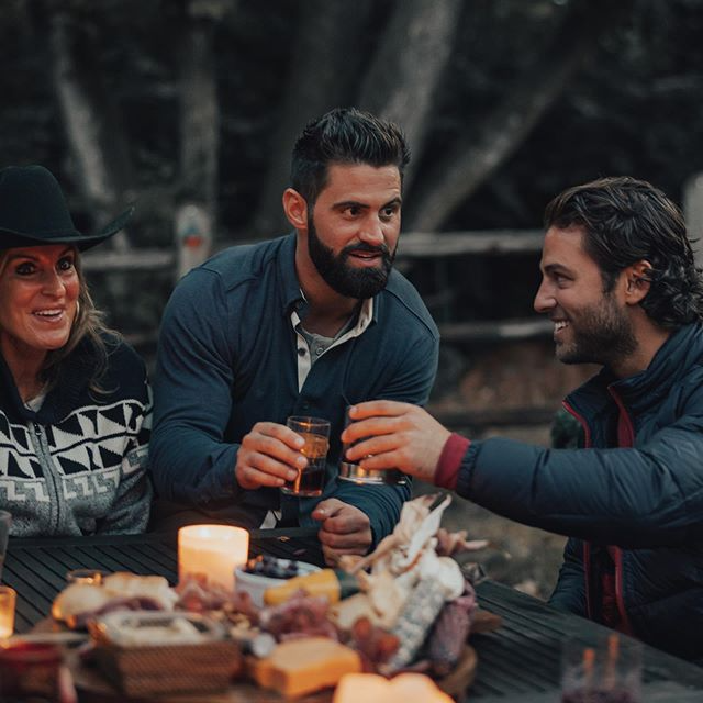Three people at a table outdoors with drinks and a charcuterie board, toasting and smiling. Evening setting with candles and a forest backdrop.