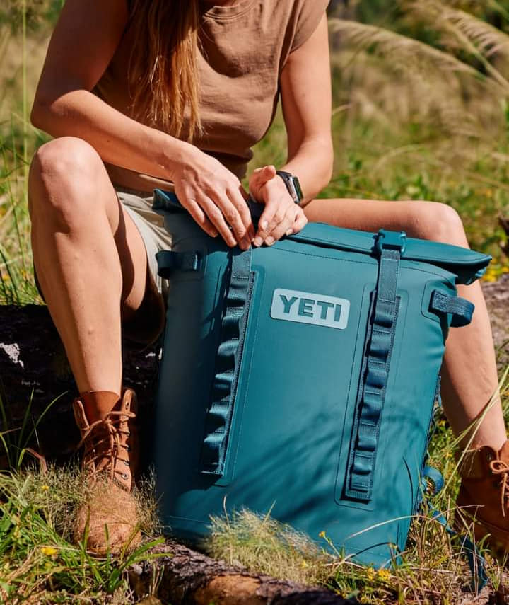 Woman opening a teal YETI backpack while sitting outdoors. She's wearing a brown shirt, shorts, and boots.