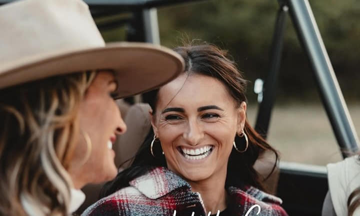 Two women in a car, laughing. One wears a plaid shirt and hoop earrings, the other a cowboy hat and large earrings. They are outside.