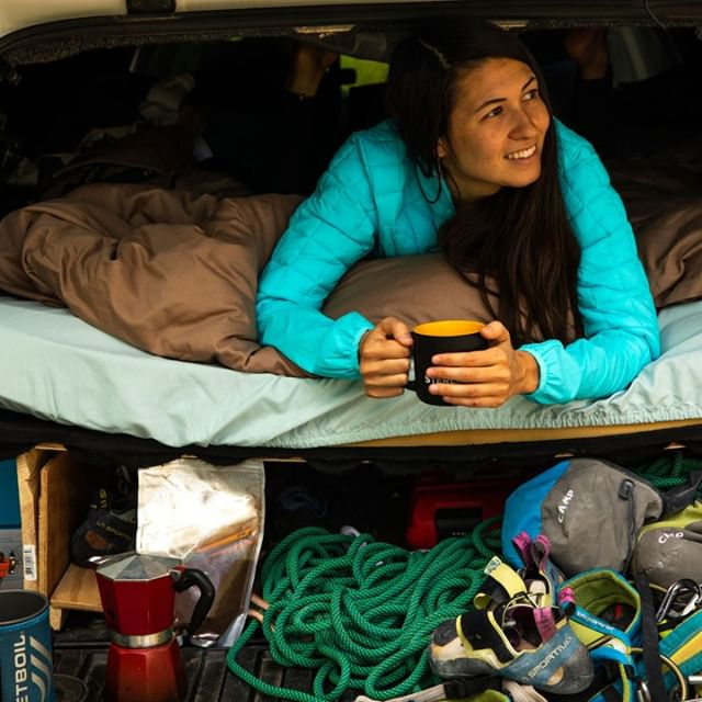 Woman in a car bed, smiling and holding a mug, surrounded by camping and climbing gear.