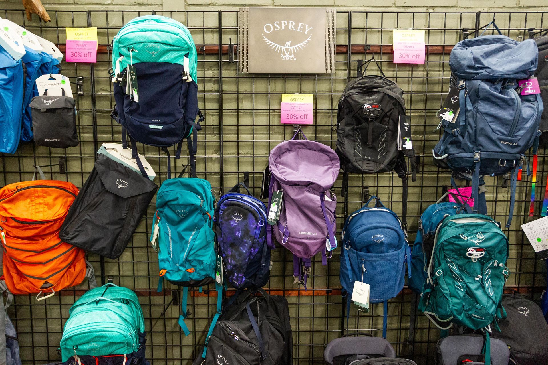 Backpacks of various colors, including blue, green, and orange, are displayed on a metal rack in a store. The Osprey brand sign is visible.