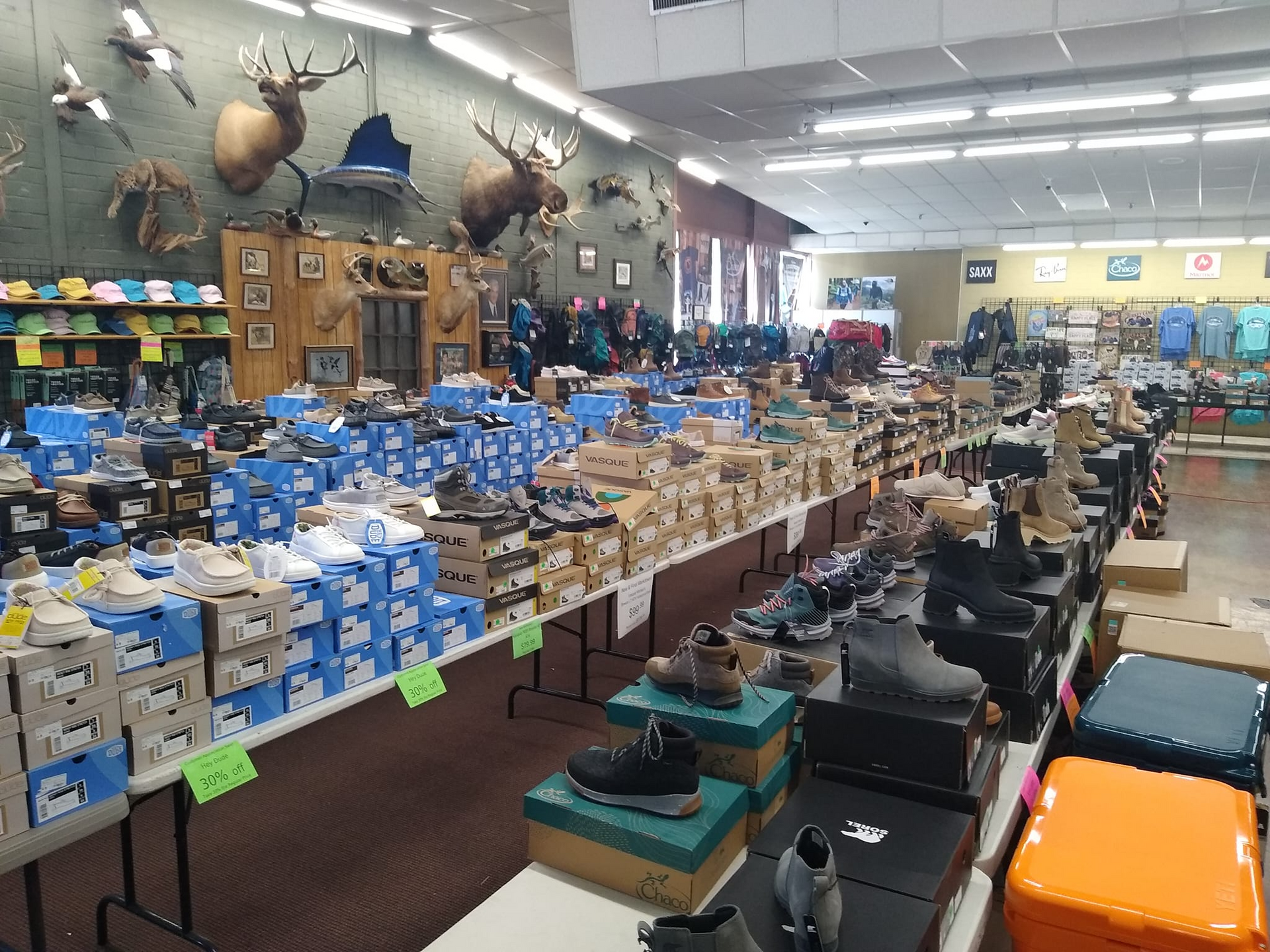 A retail store displaying various shoes, including hiking boots, on tables. Wildlife mounts and outdoor gear are visible in the background.