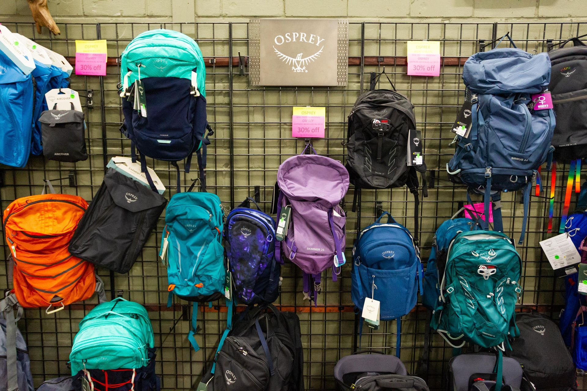 Display of colorful Osprey backpacks on a store wall. Various sizes and colors like turquoise, purple, and orange are shown.