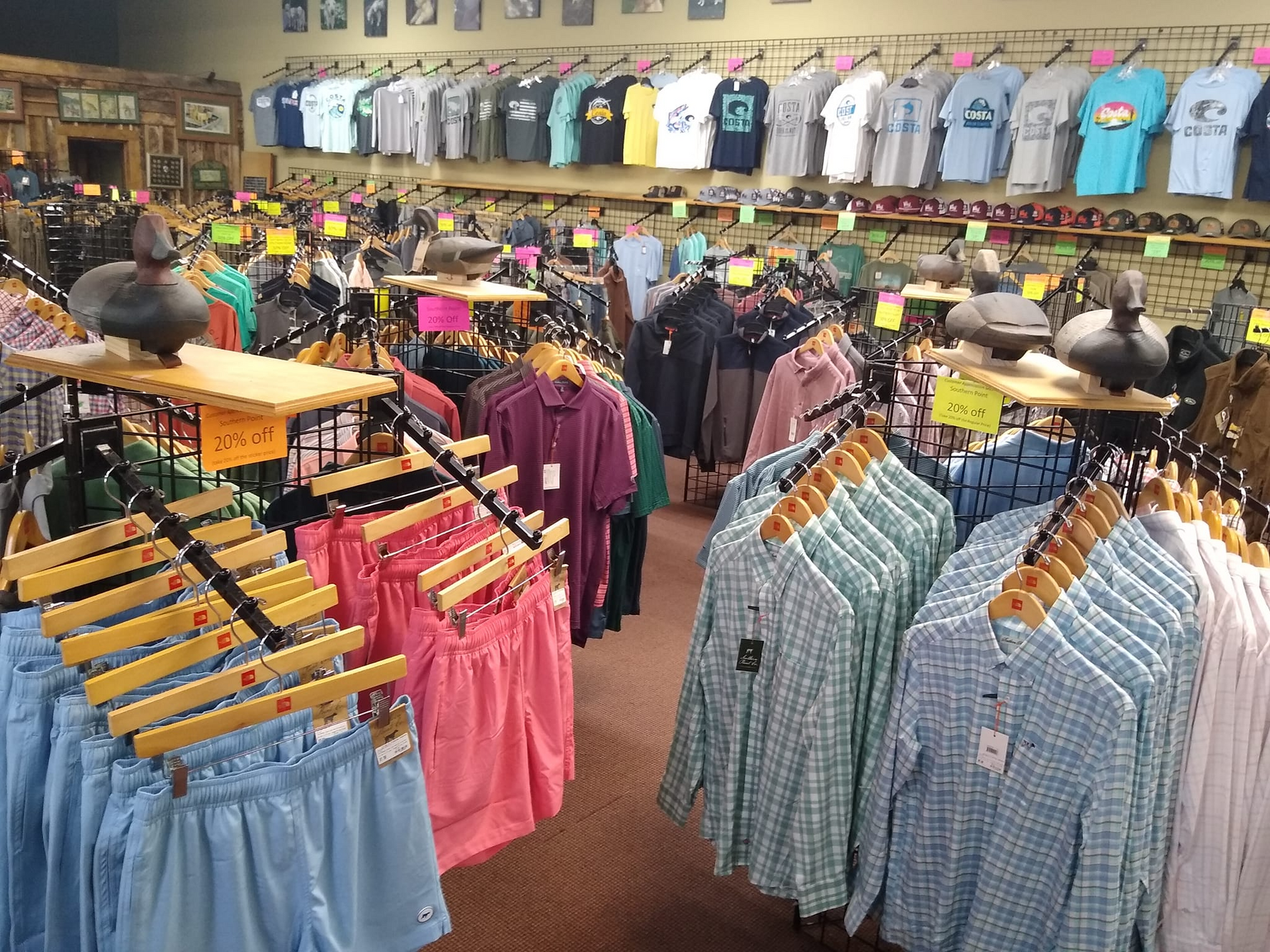 Inside a retail store, clothing racks display shorts and shirts. Duck decoys sit on shelves.