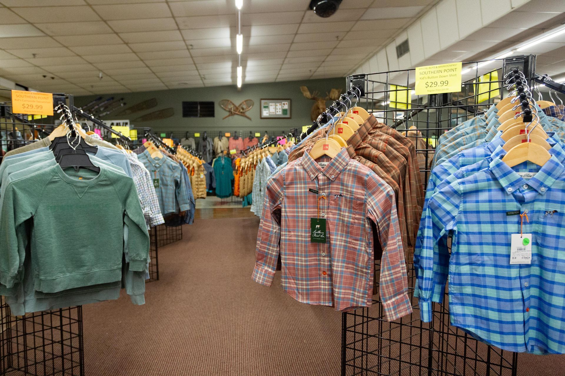 Clothing racks in a store, displaying children's shirts and sweatshirts. The store has a wood-paneled floor and fluorescent lighting.