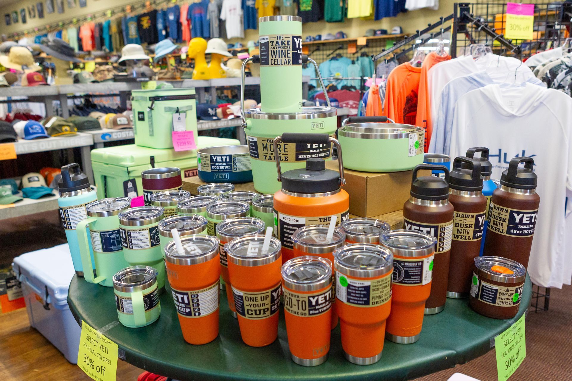 Display of orange and teal YETI drinkware in a retail store, with coolers, clothing, and hats in the background.