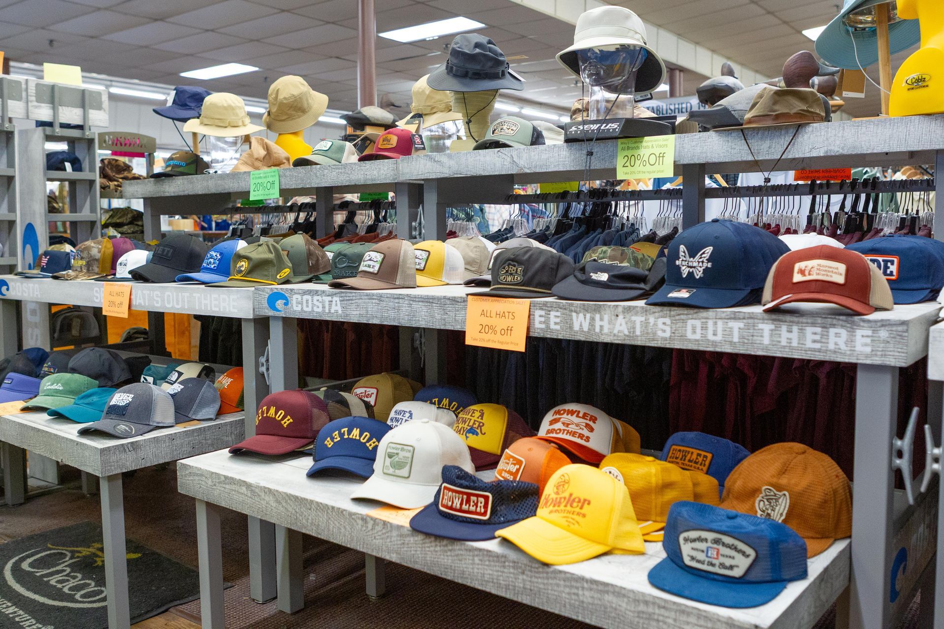 A store display with rows of hats in various colors, including baseball caps and bucket hats. The shelves have signs and are gray.