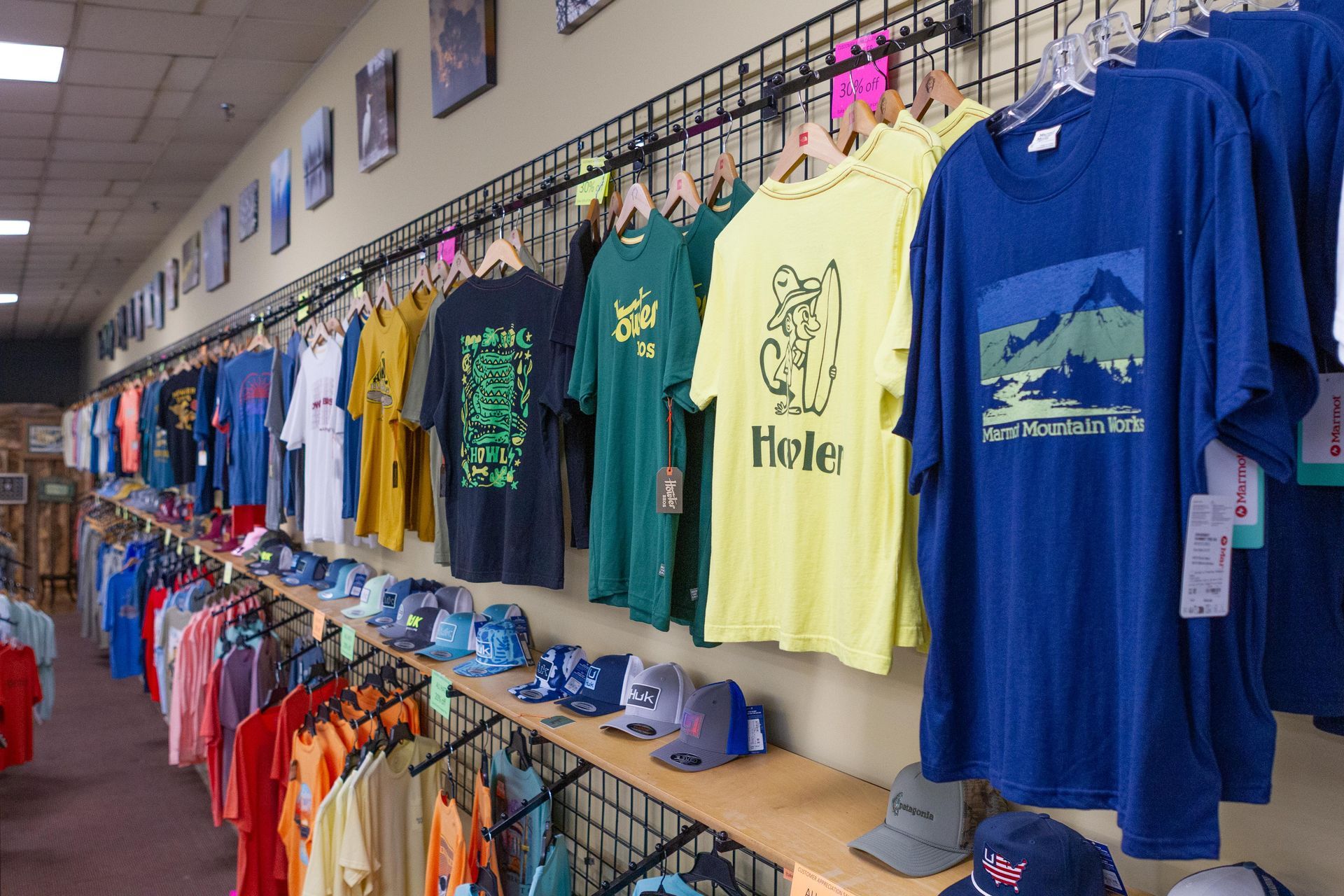 Inside a retail store, a display of colorful t-shirts hang on racks, with hats and more shirts on shelves below.