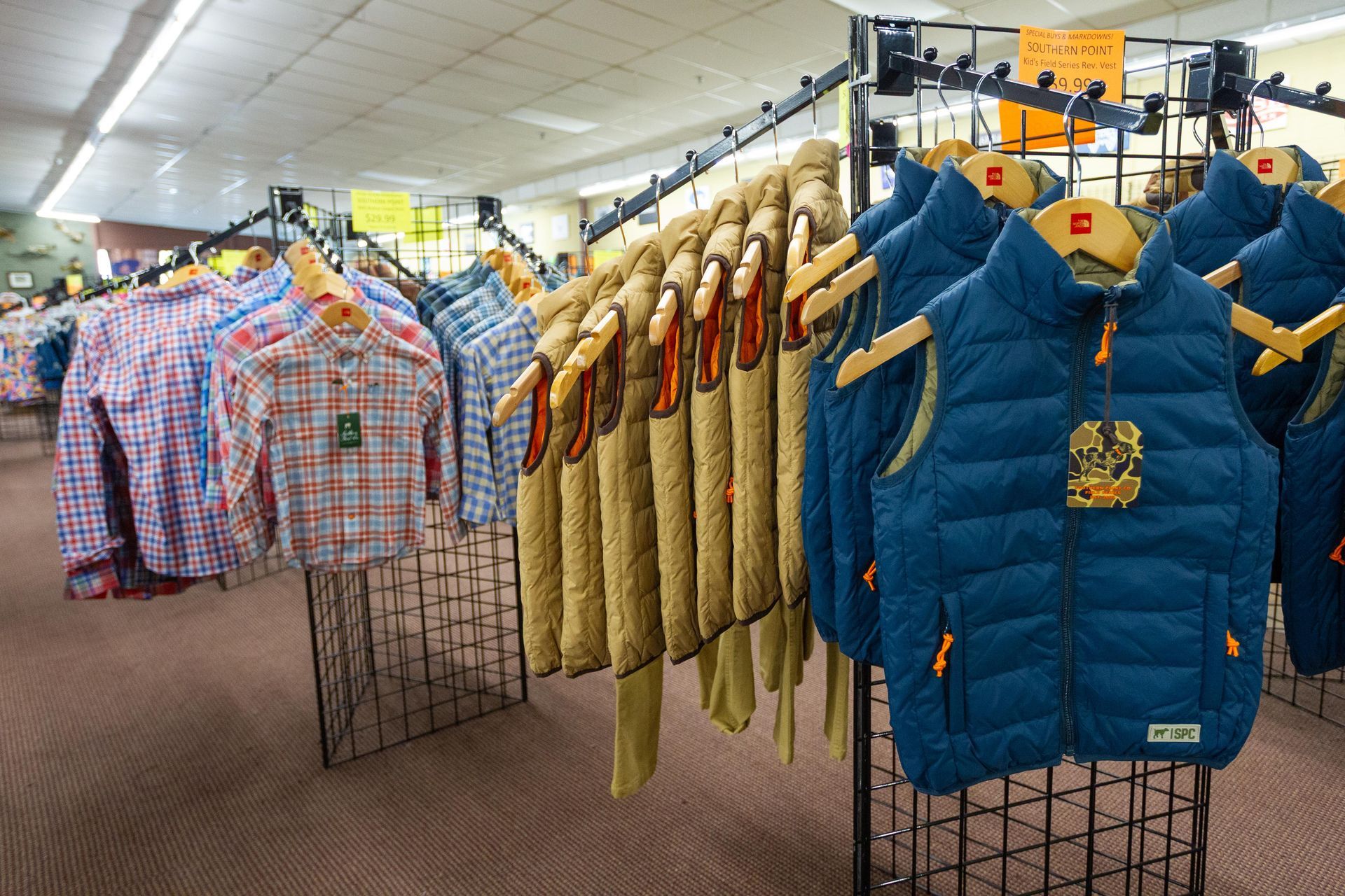 Clothing racks in a store display plaid shirts, knit vests, and blue puffer vests. The store has black racks on a carpeted floor.