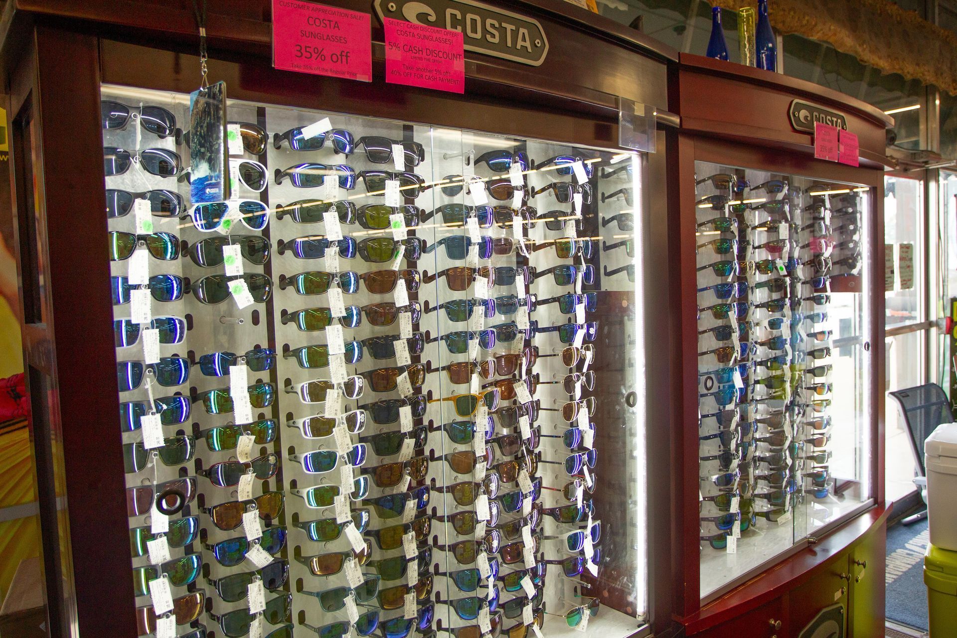 Sunglasses displayed in a wooden cabinet at a shop. Many styles and colors are visible, arranged in rows.