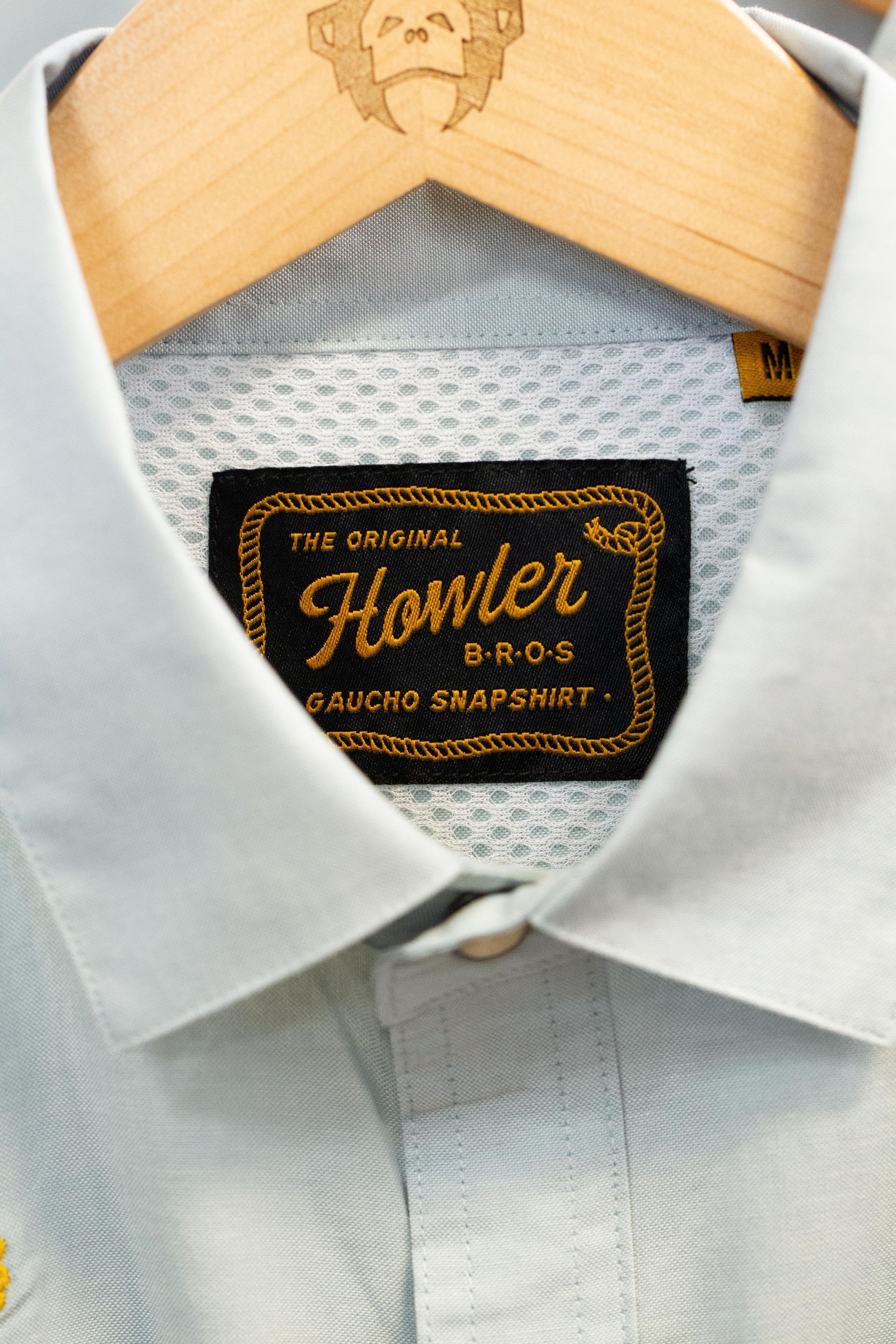 Close-up of a Howler Bros shirt collar with the brand label. Light blue shirt with a textured collar, wood hanger.