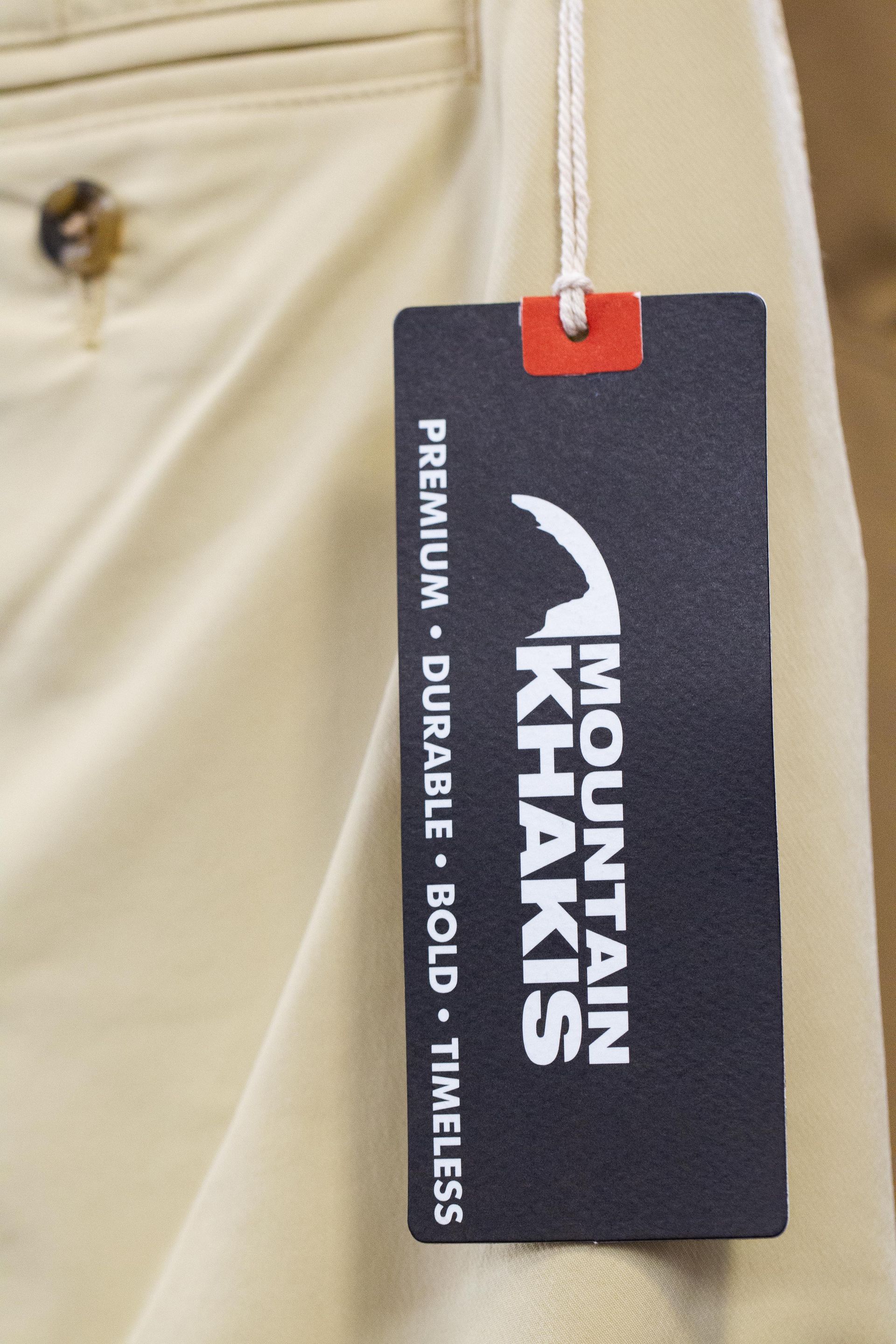 A black Mountain Khakis tag hangs from beige pants. The tag reads 