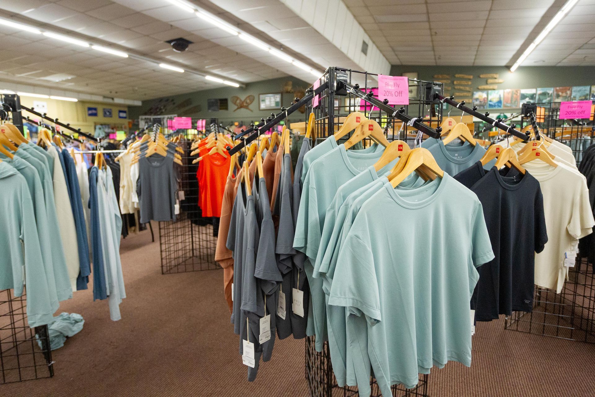 Clothing racks filled with t-shirts and other apparel in a store setting. Clothes are various colors, with white, blue, and orange shirts visible.