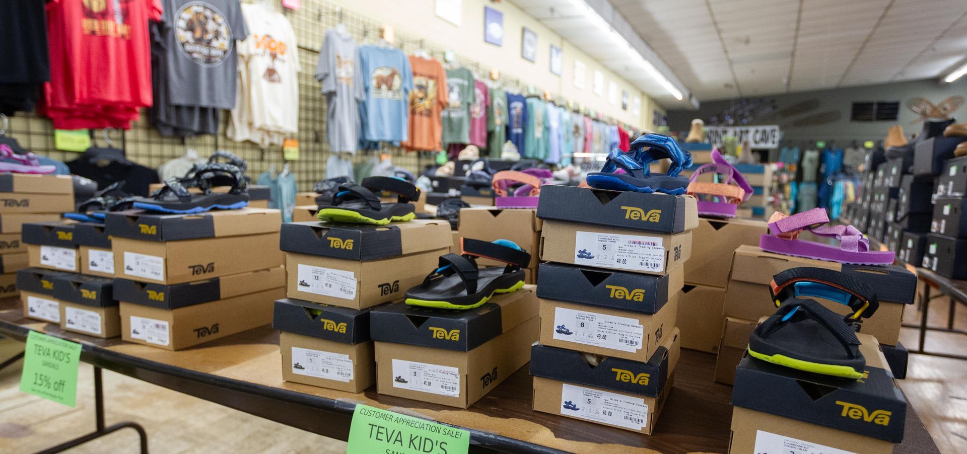 A store display showcasing stacks of Teva sandals in boxes on a table, with clothing racks and other merchandise in the background.