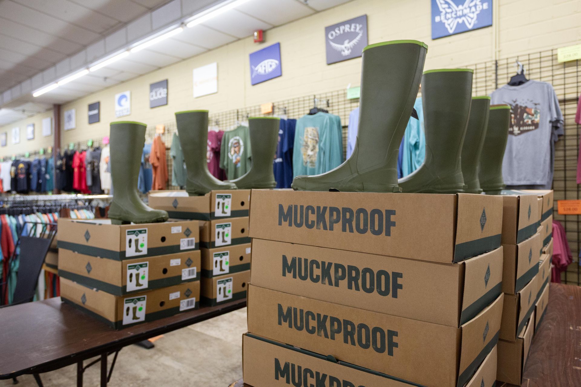 Green MuckProof boots on display atop stacked boxes in a store, with clothing racks and signs in the background.