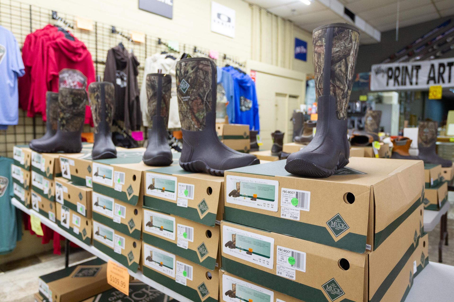 Boots for sale displayed on boxes in a store, with camouflage design and various sizes. Behind the boots, clothing hangs on a rack.