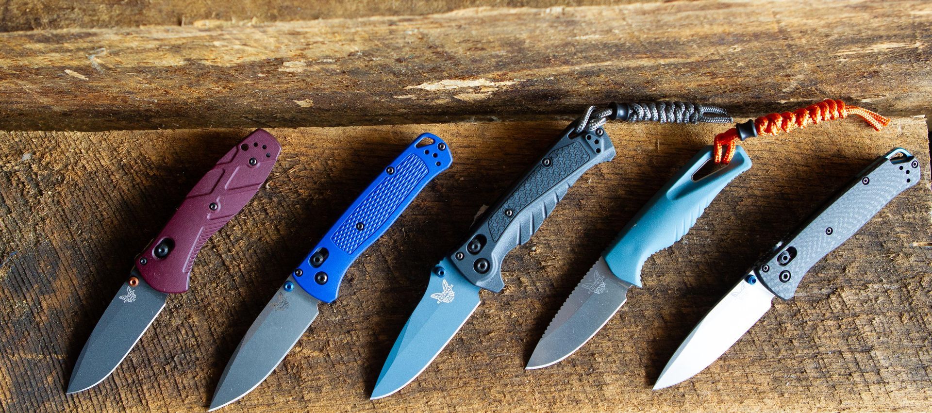 Five folding knives with different colored handles are arranged on a wooden surface.