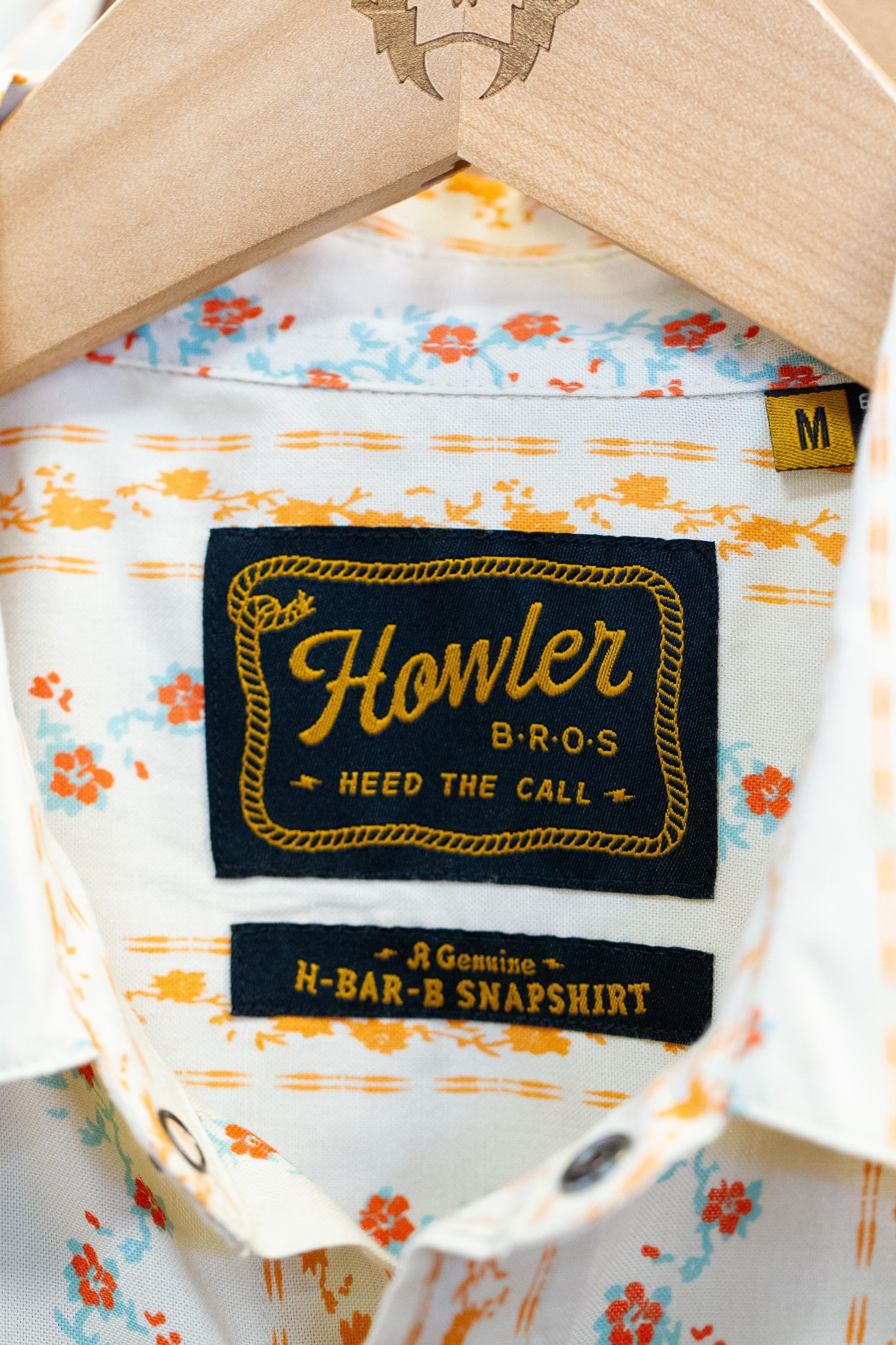 Close-up of a Howler Bros shirt tag on a wooden hanger. The tag is black with gold lettering and a floral pattern background.