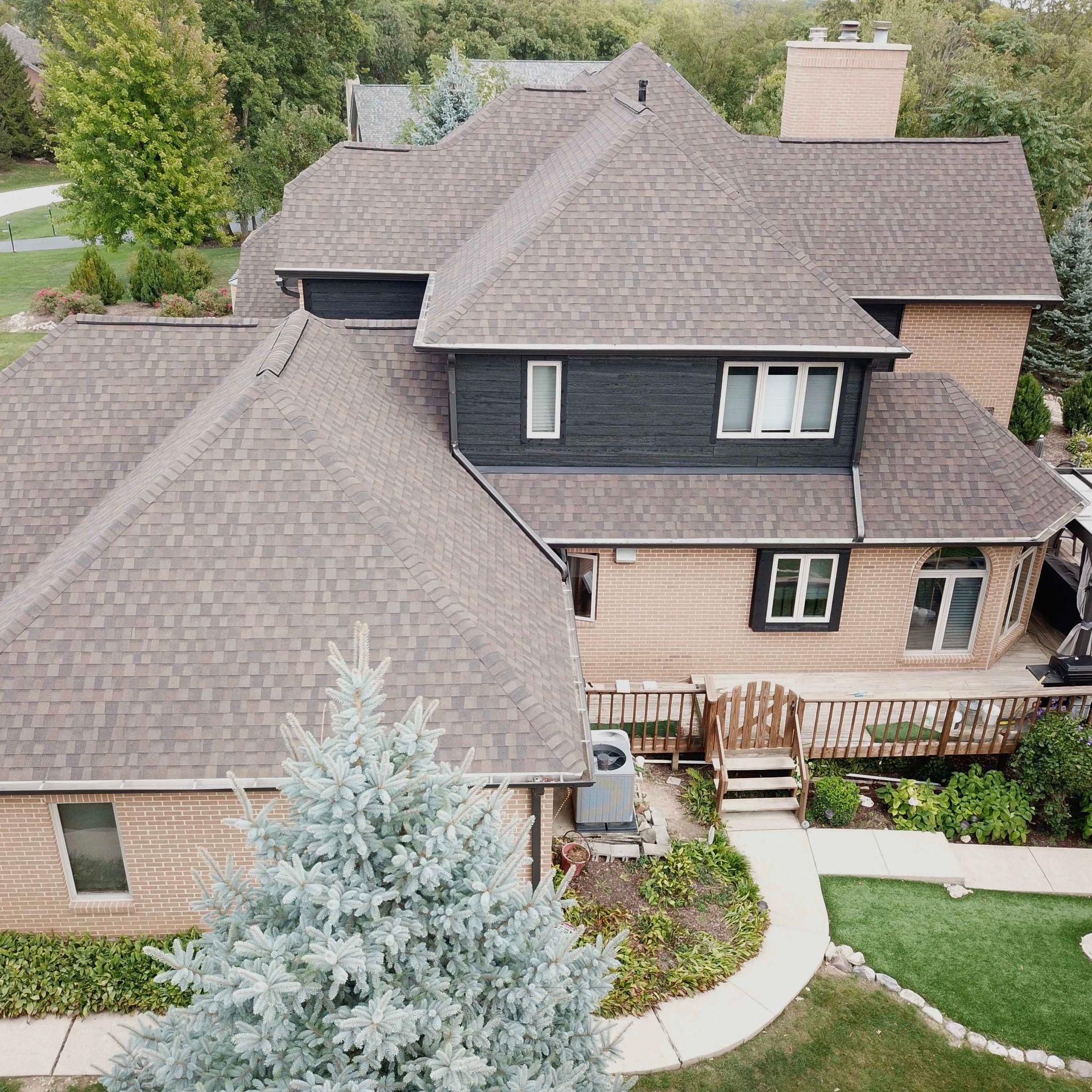 Aerial view of a large house with multiple roof sections. Light brown shingles, wooden siding, and a deck are visible.