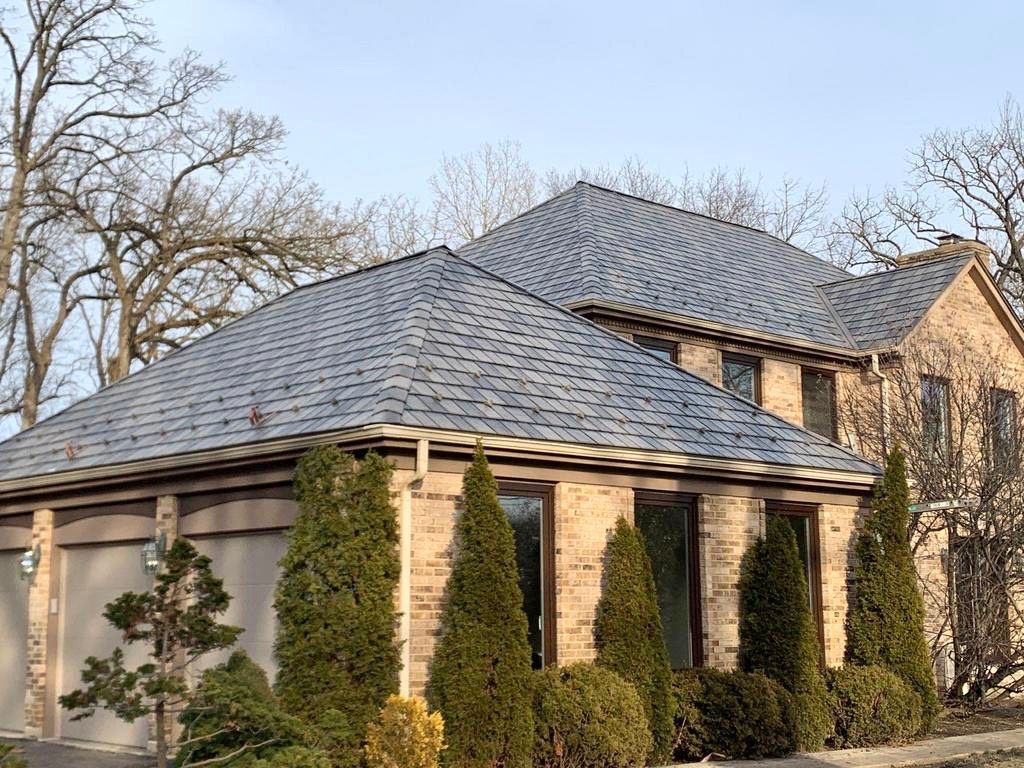 Beige brick house with dark gray shingle roof, green bushes, and bare trees against a blue sky.