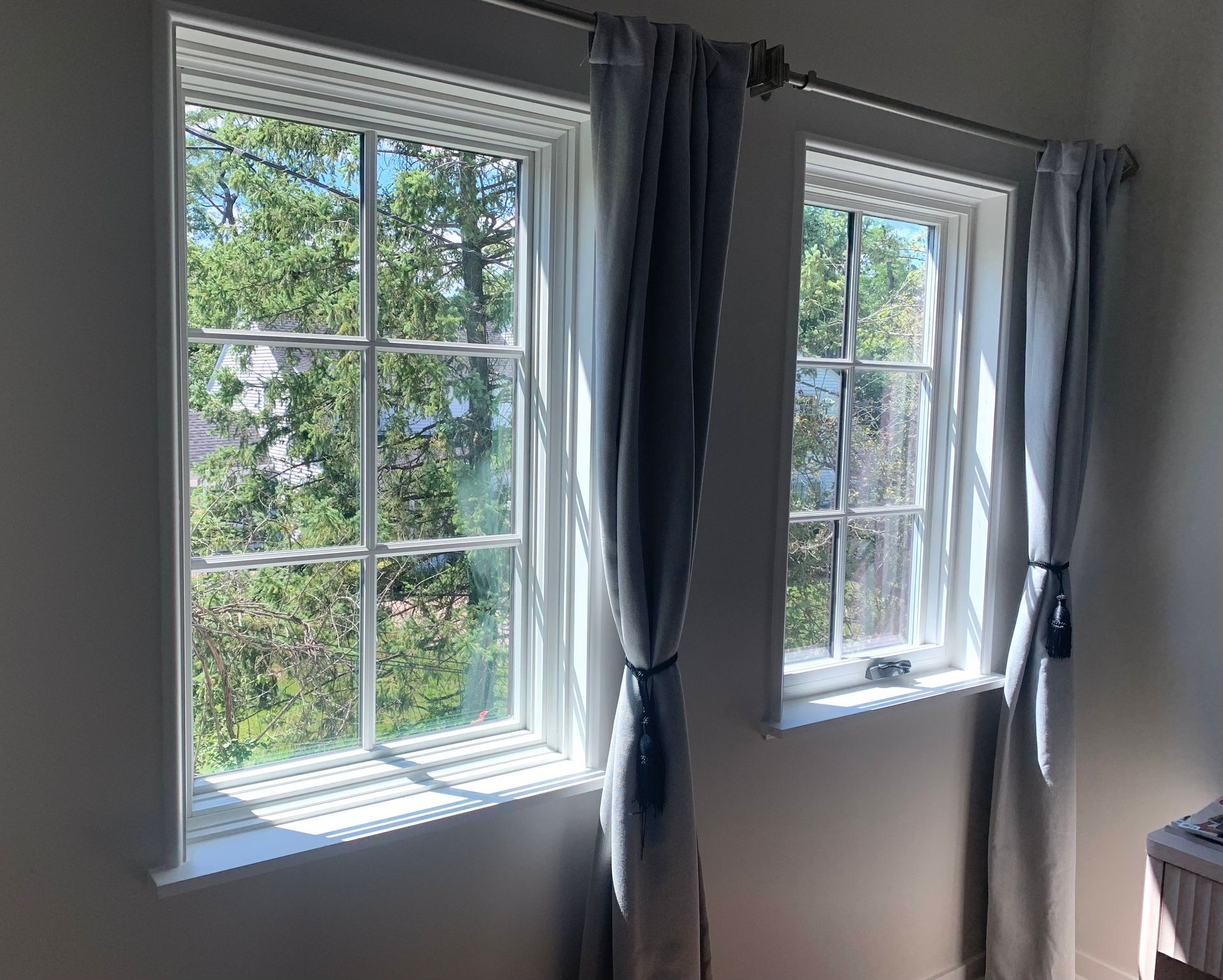 Two white-framed windows with light gray curtains. Outside is a view of green trees and sky. Sunlight streams in.