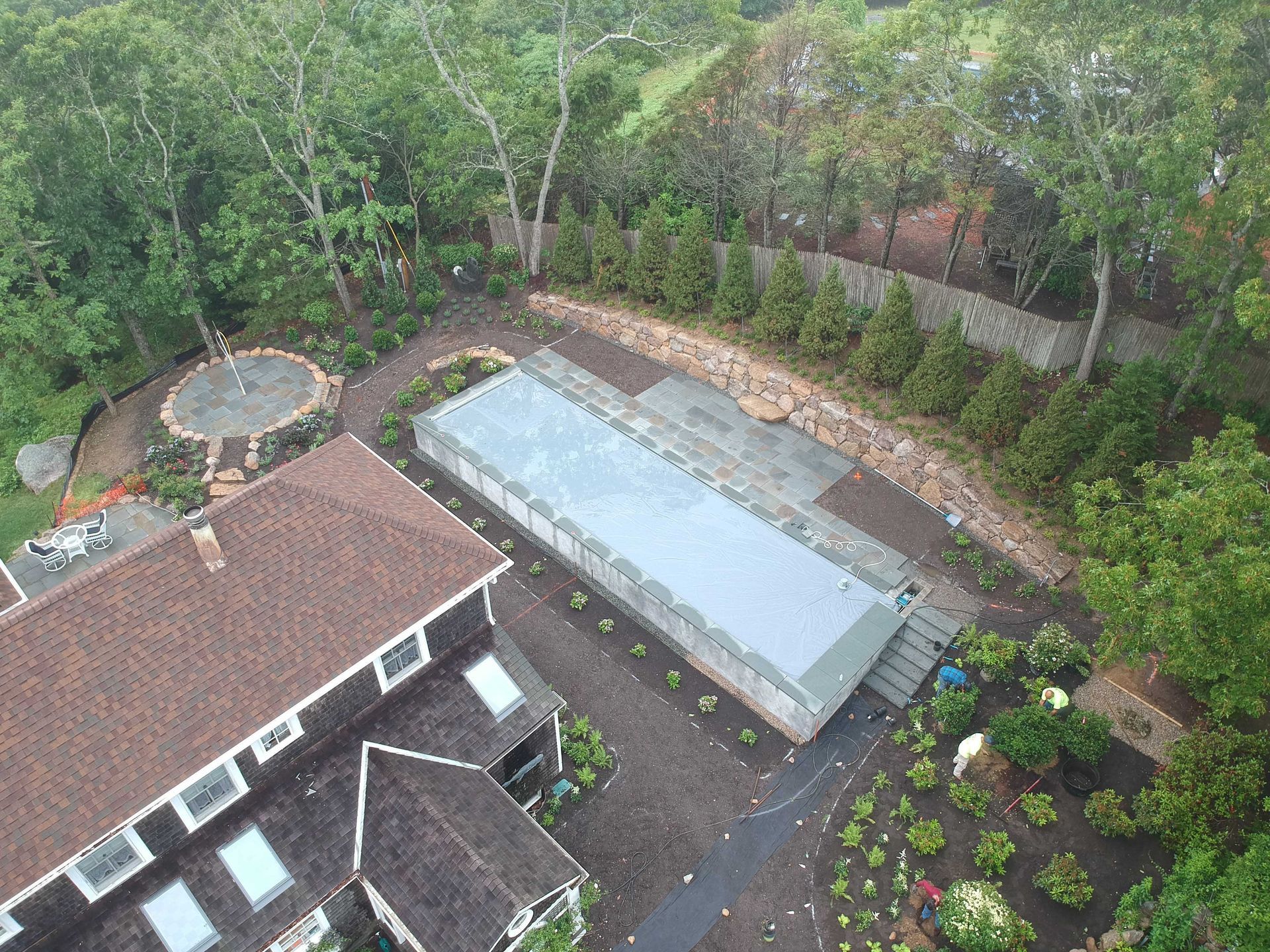 Aerial view of a house with a covered rectangular pool, stone patio, and landscaped yard in a wooded setting.