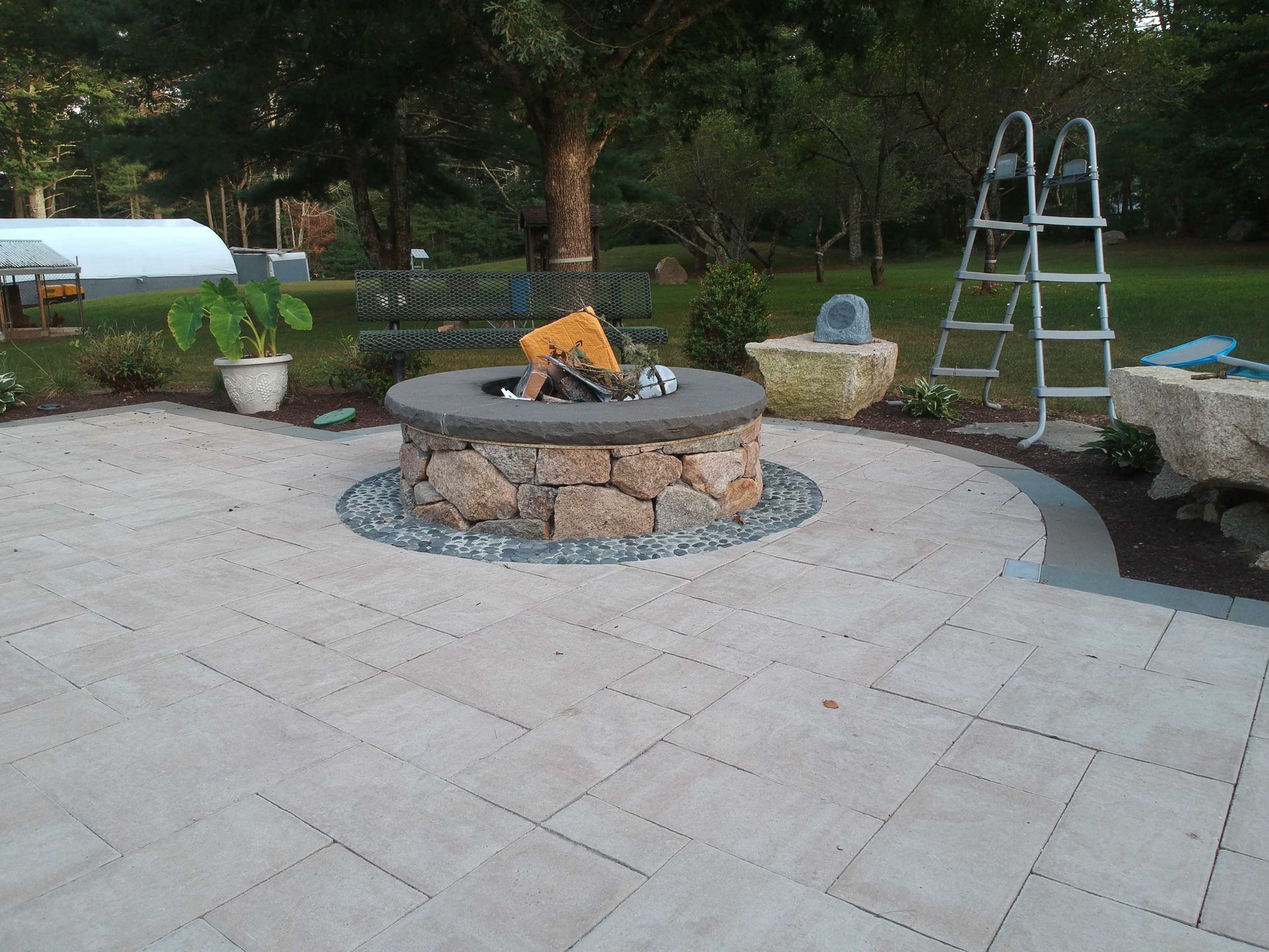 A round stone fire pit sits on a light-colored paver patio, with a metal ladder and garden decor nearby in a grassy yard.