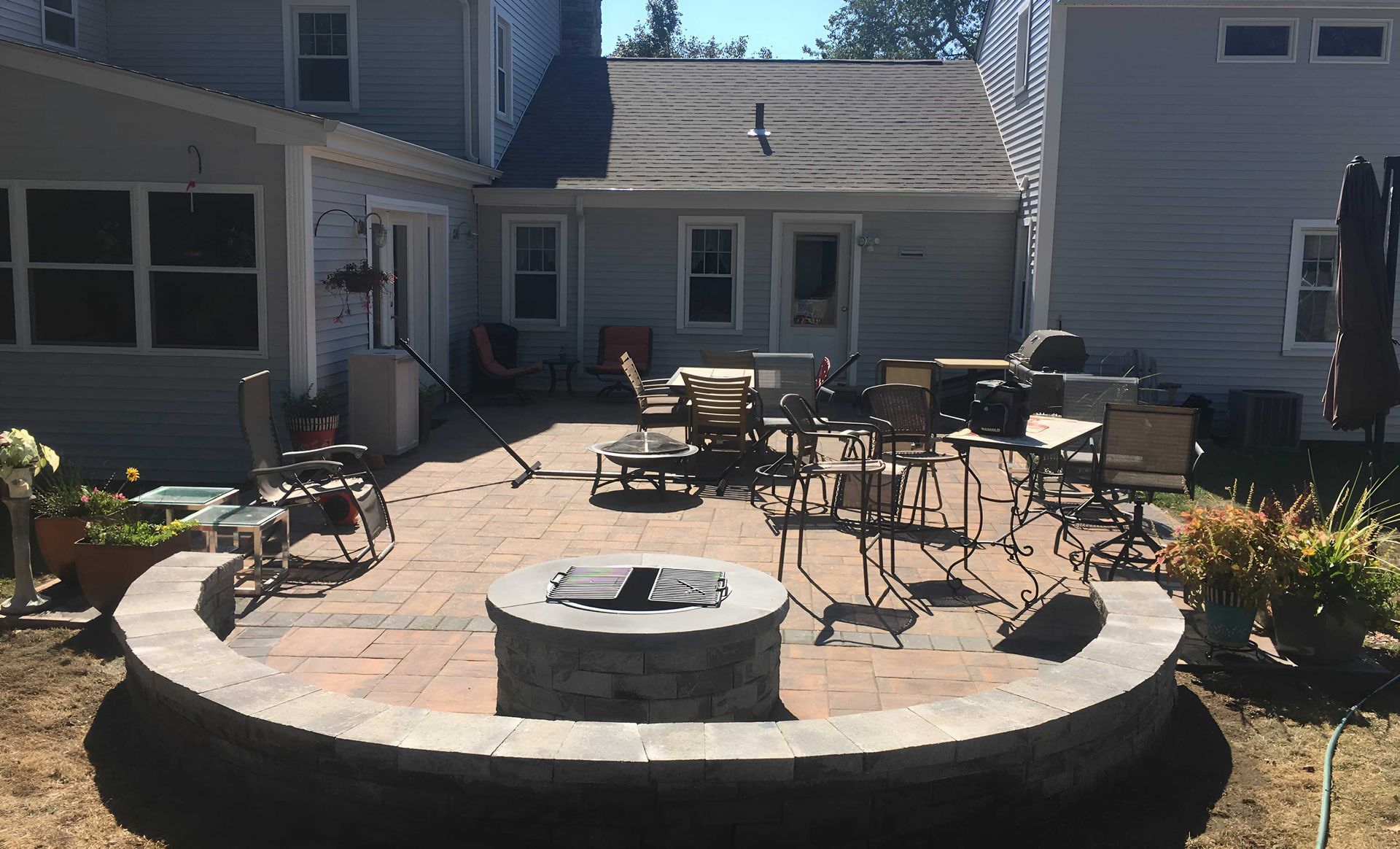 A stone patio features a central fire pit surrounded by a curved retaining wall, outdoor chairs, and dining furniture.