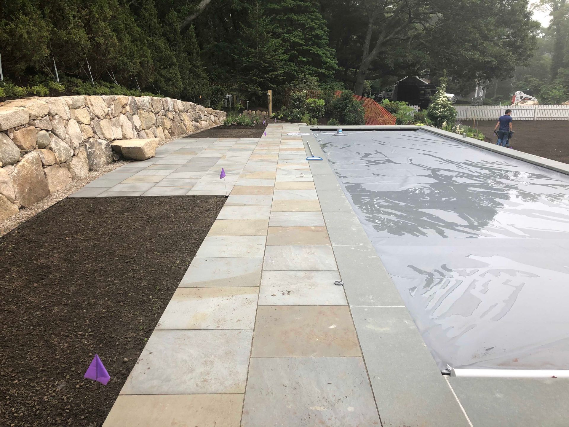 A stone patio borders a covered swimming pool next to a large stone retaining wall and areas of dark soil.