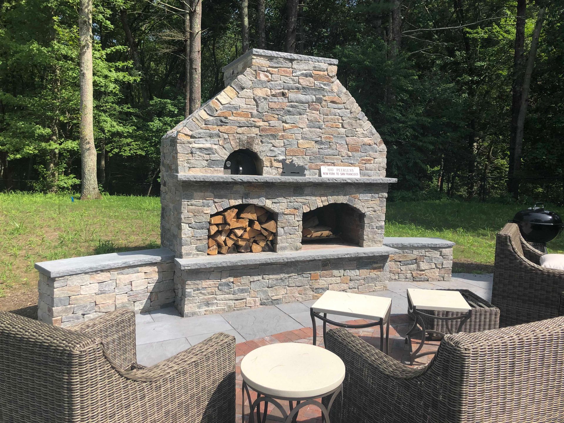 A stone outdoor fireplace with a wood-burning pizza oven, firewood storage, and a patio seating area in a wooded yard.