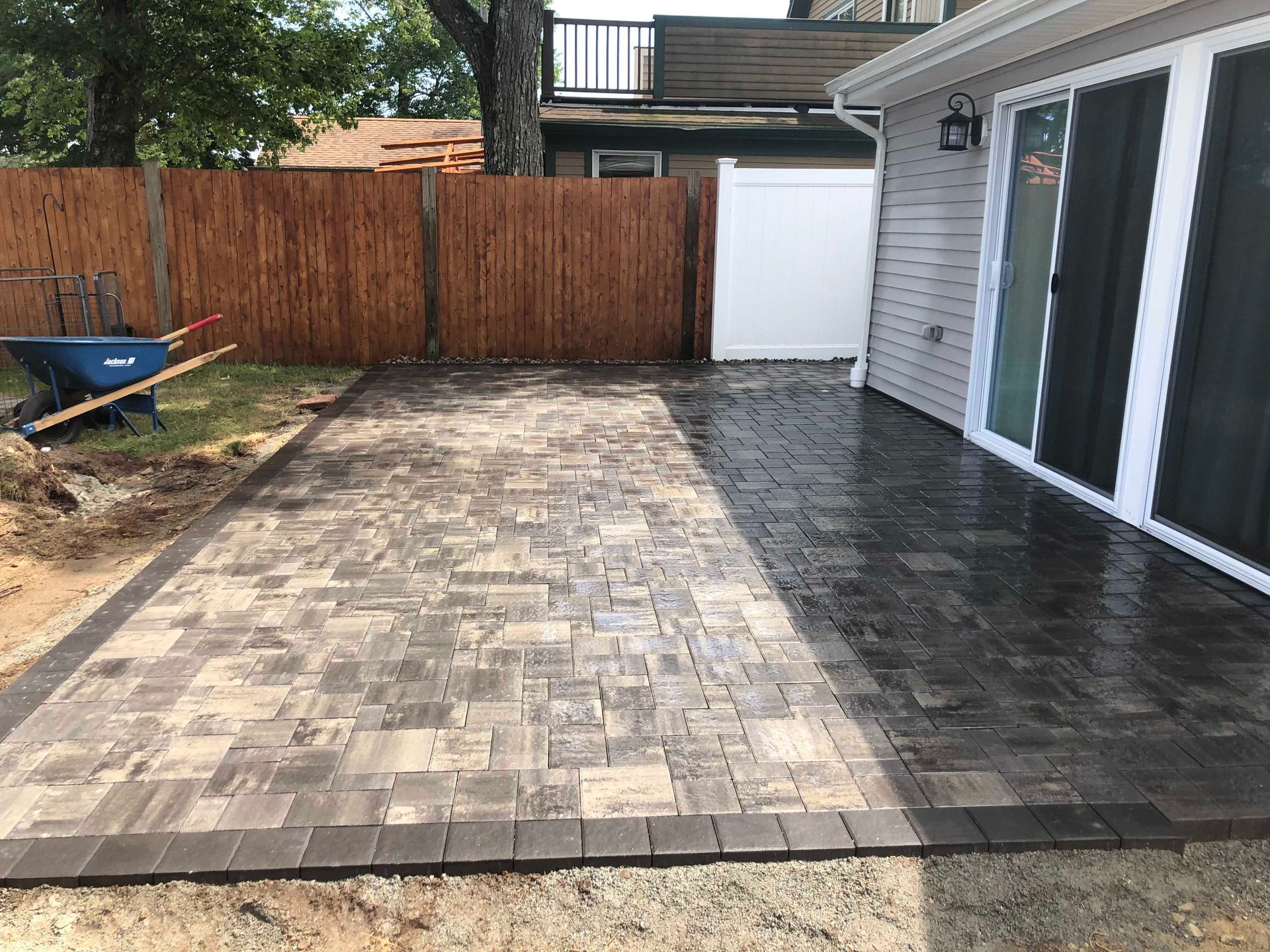 A patio made of two types of pavers—light gray and dark gray—next to a house with a sliding door and a wooden fence nearby.