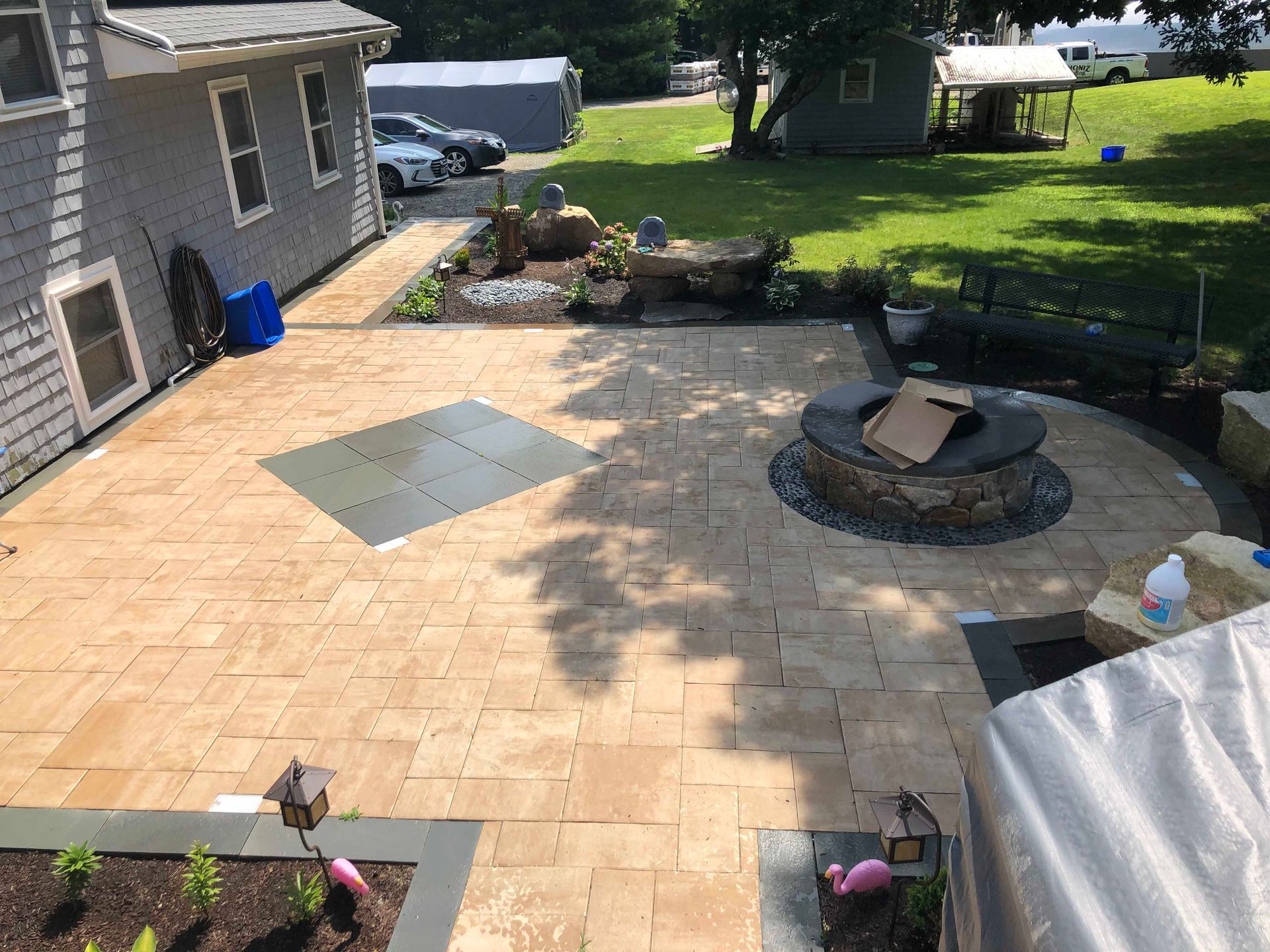 A tan paver patio with a circular stone fire pit, a square stone inlay, and a grassy backyard.