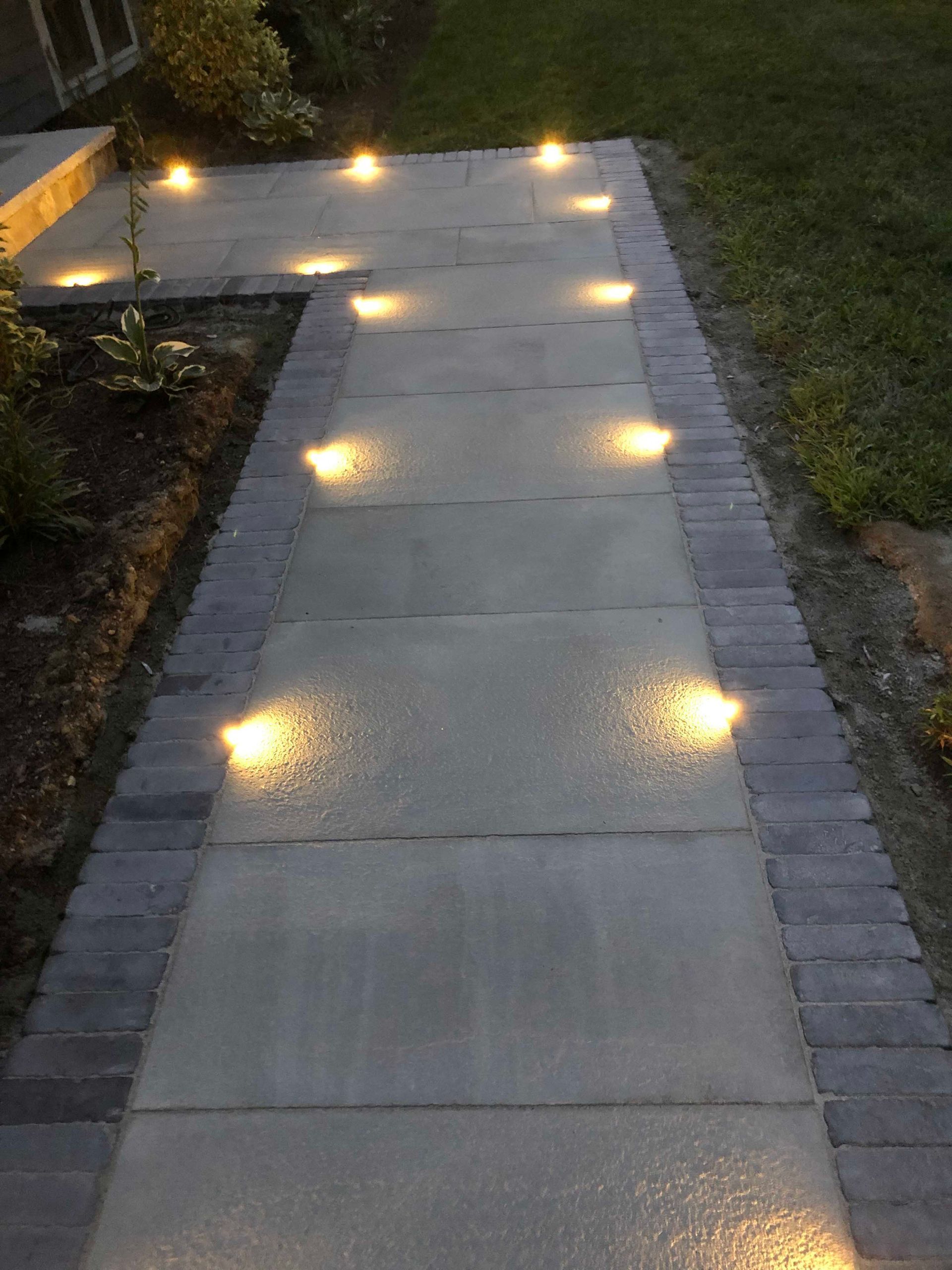 A paved stone walkway at twilight, illuminated by recessed warm-light LED ground lights along both edges.