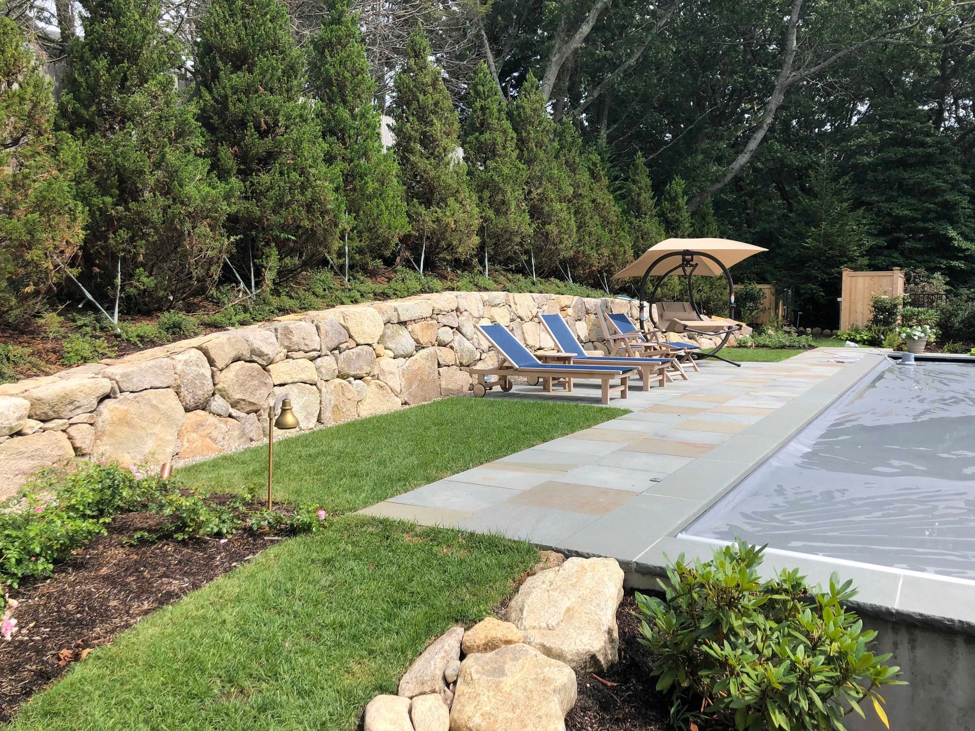 A stone retaining wall overlooks a swimming pool surrounded by lounge chairs, a patio, and a line of tall evergreen trees.