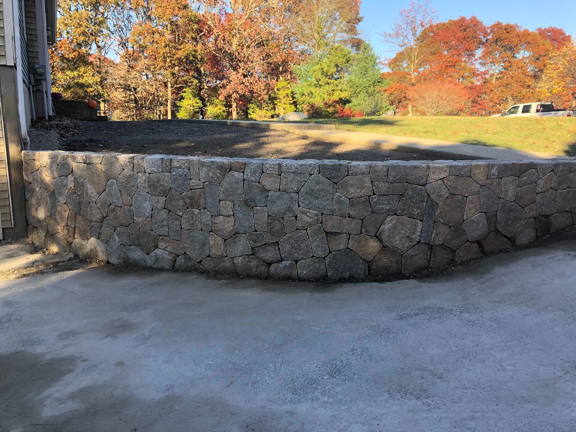 A curved, mortared stone retaining wall stands in a yard with autumn trees in the background under a clear blue sky.