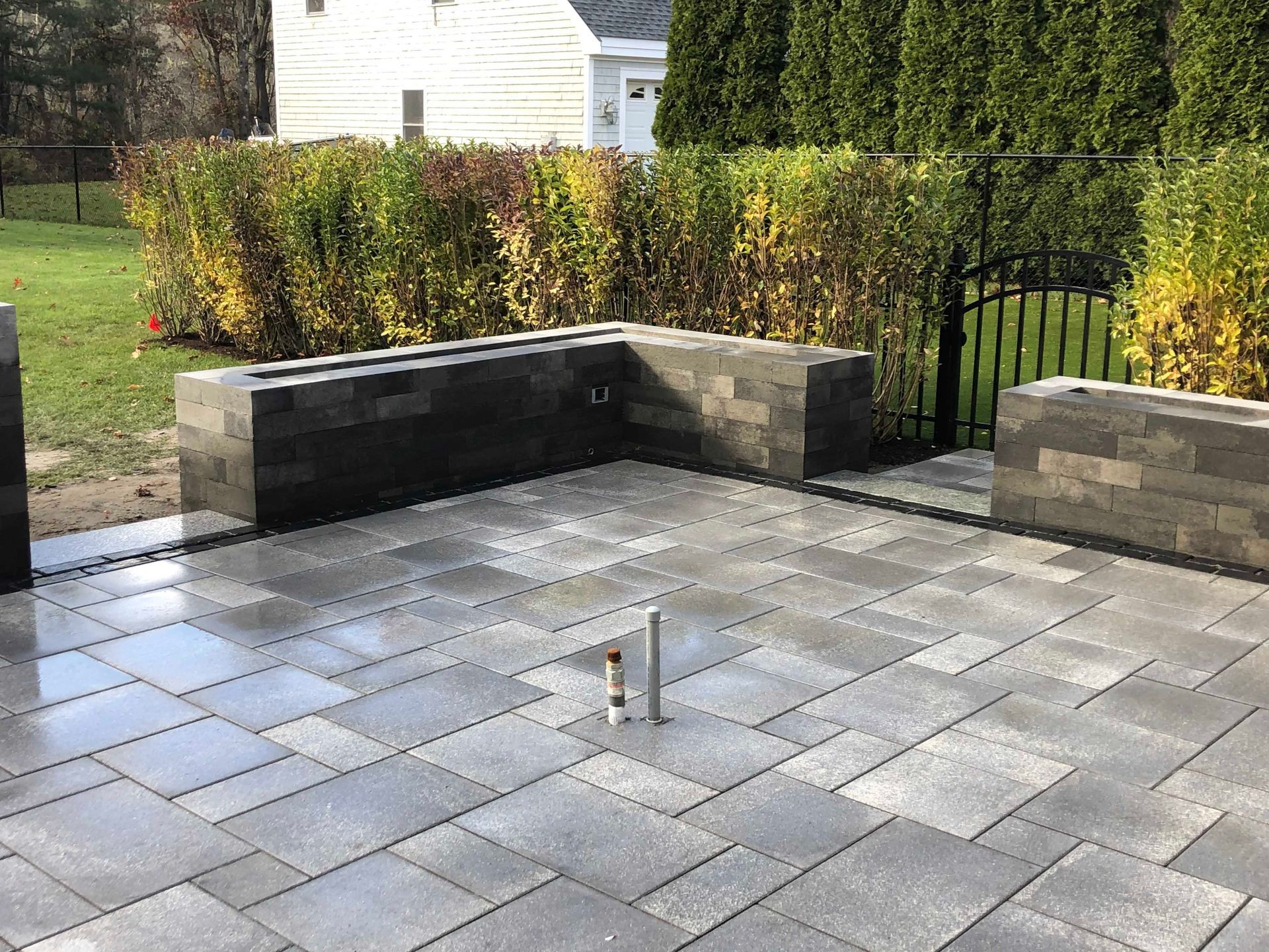A gray paver patio with a low L-shaped stone retaining wall and a small pipe protrusion, set against a hedge and house.