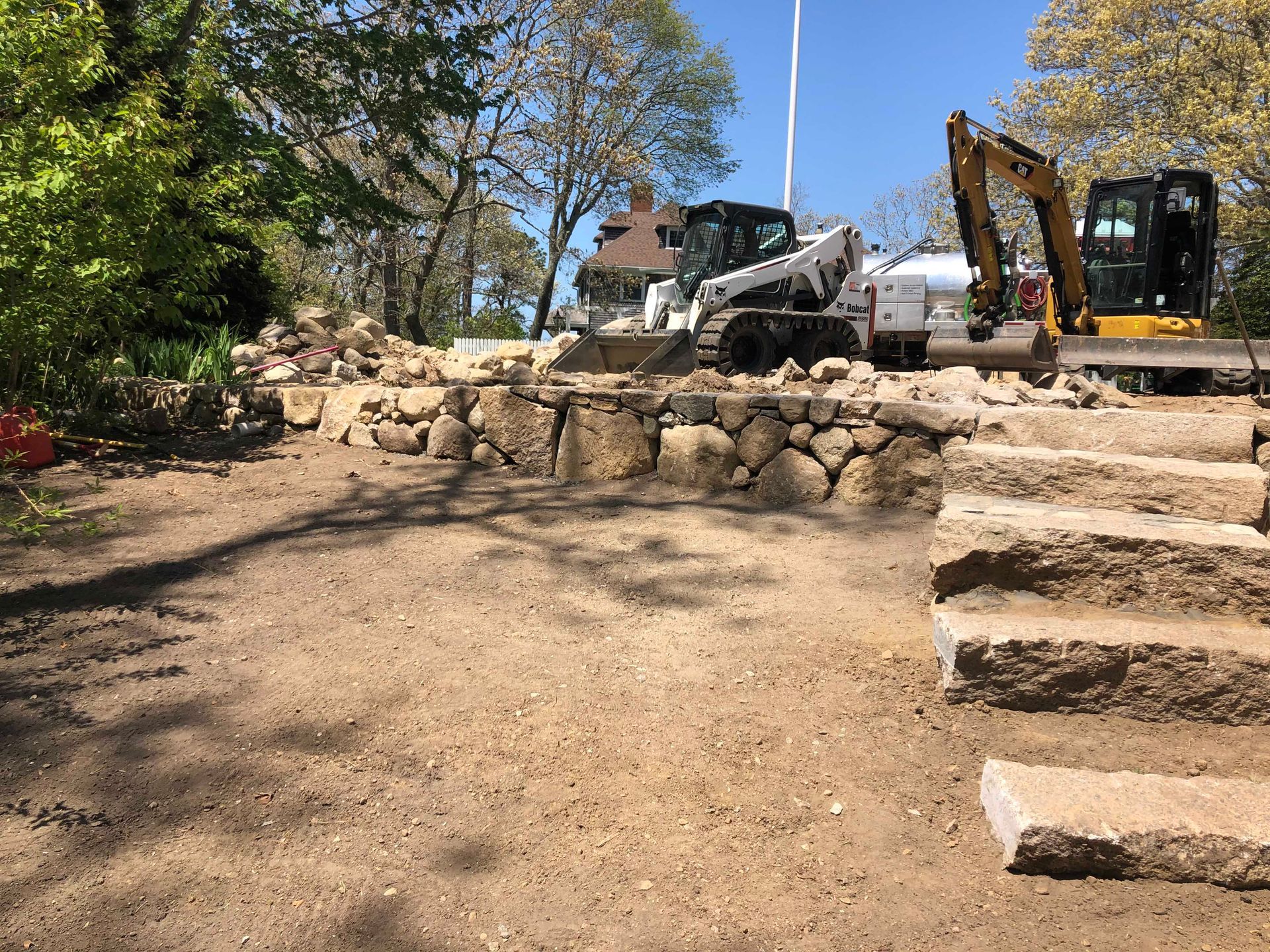 A skid-steer and an excavator parked above a stone retaining wall and stone steps under construction in a sunny yard.