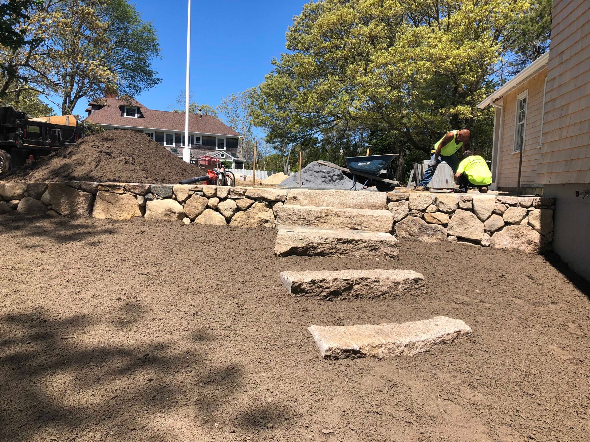 Workers in high-visibility vests finish a stone retaining wall and landscape stairs in a yard under a clear blue sky.