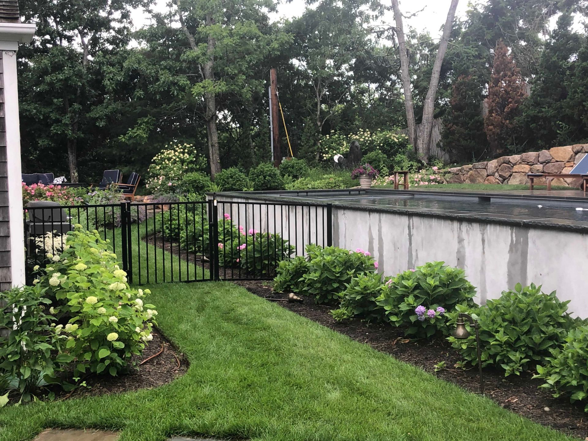A grassy path leads to a black metal fence beside a large concrete retaining wall, surrounded by lush green shrubs.