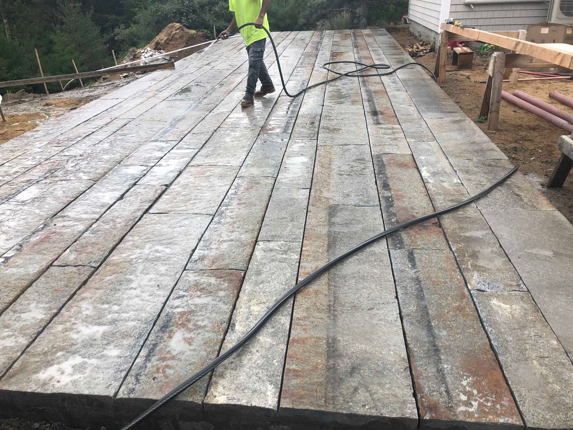A worker in a neon yellow shirt uses a hose to spray a large patio surface made of long, rectangular stone slabs.