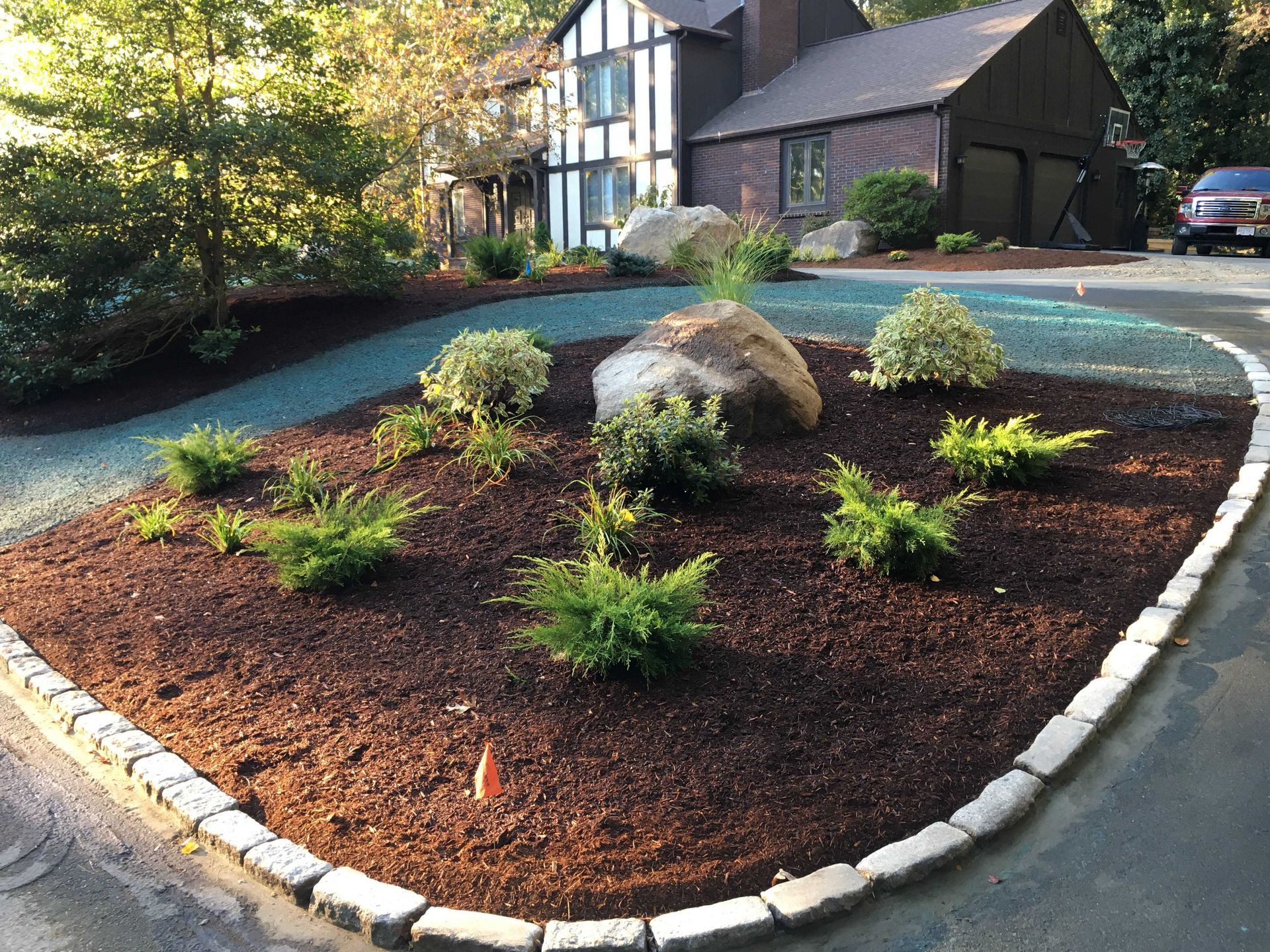 A landscaped garden bed filled with dark mulch, shrubs, and a central boulder, edged by light-colored stone bricks.