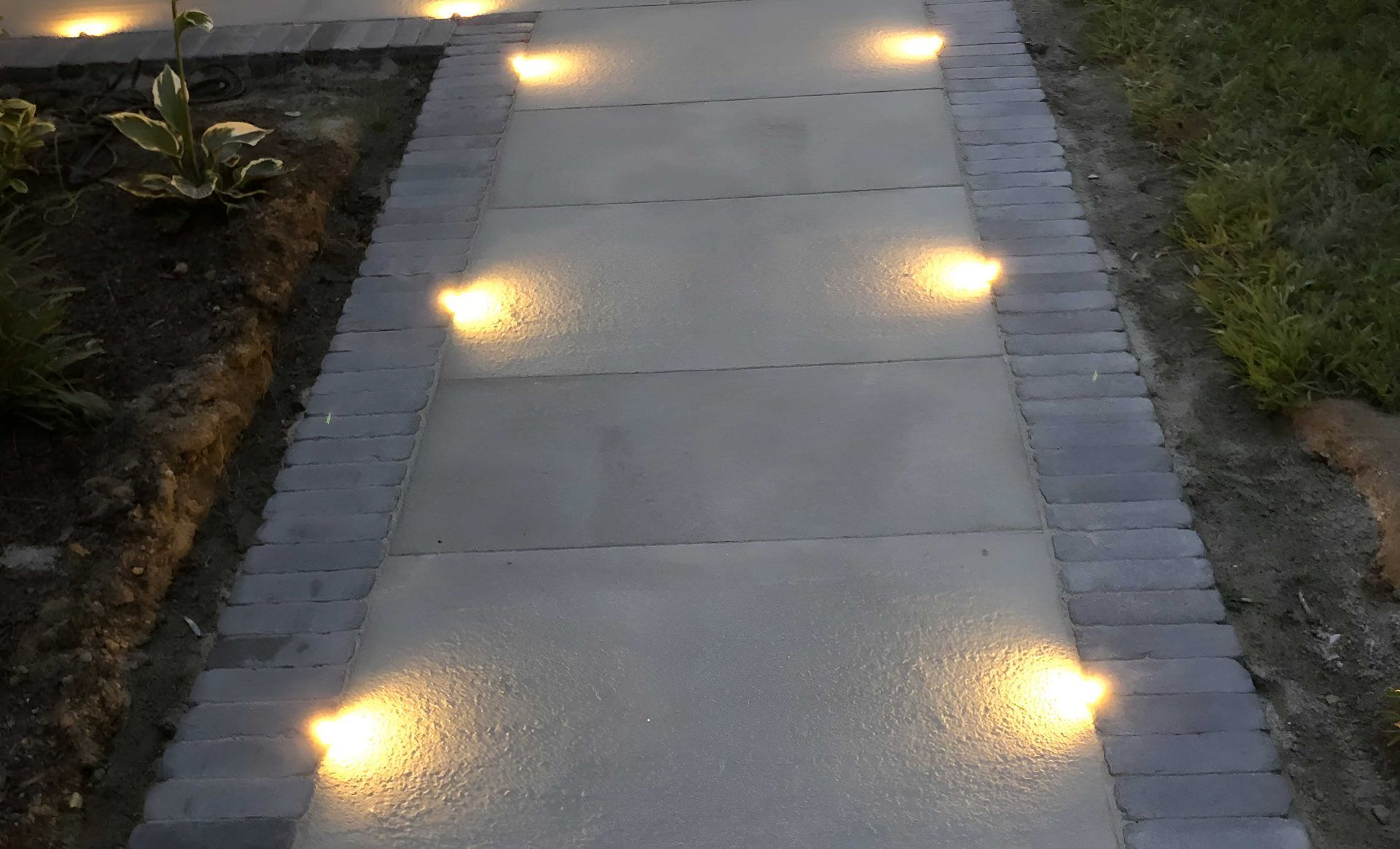 A paved walkway at night with warm, circular ground-level lights embedded along both sides.