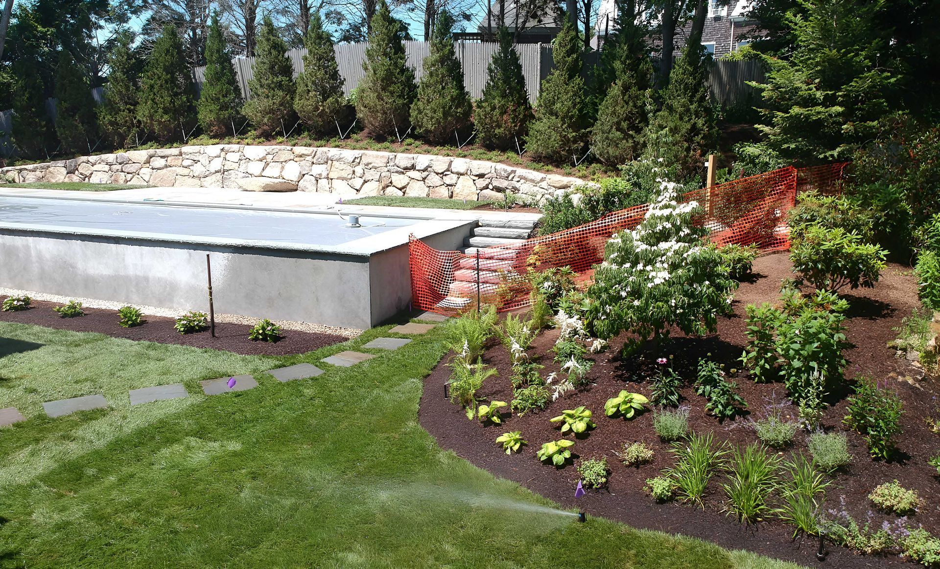 A backyard featuring a pool with a grey cover, a stone retaining wall, evergreen trees, and a landscaped garden bed.
