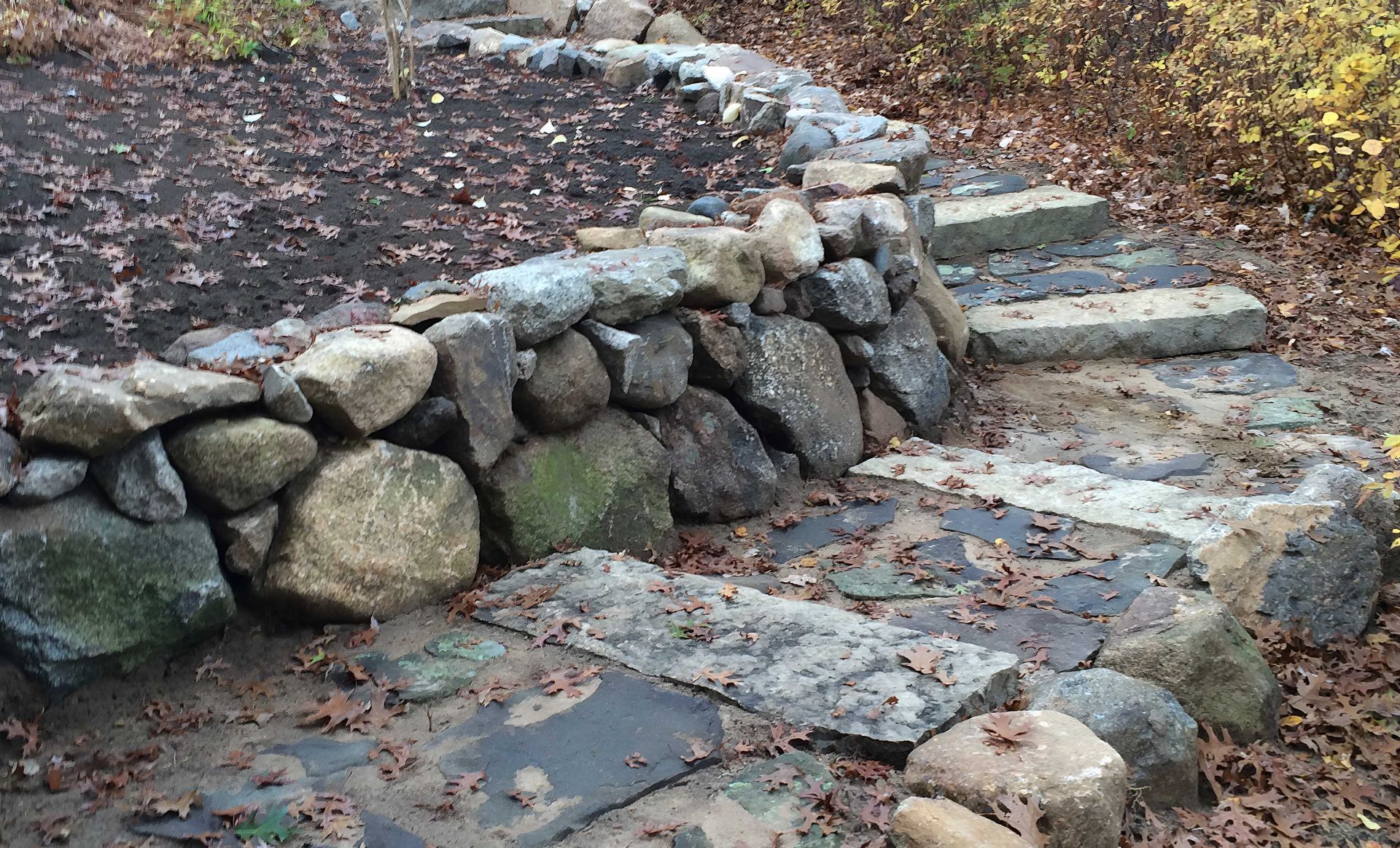 A stone retaining wall curves beside a set of stone steps surrounded by fallen autumn leaves.