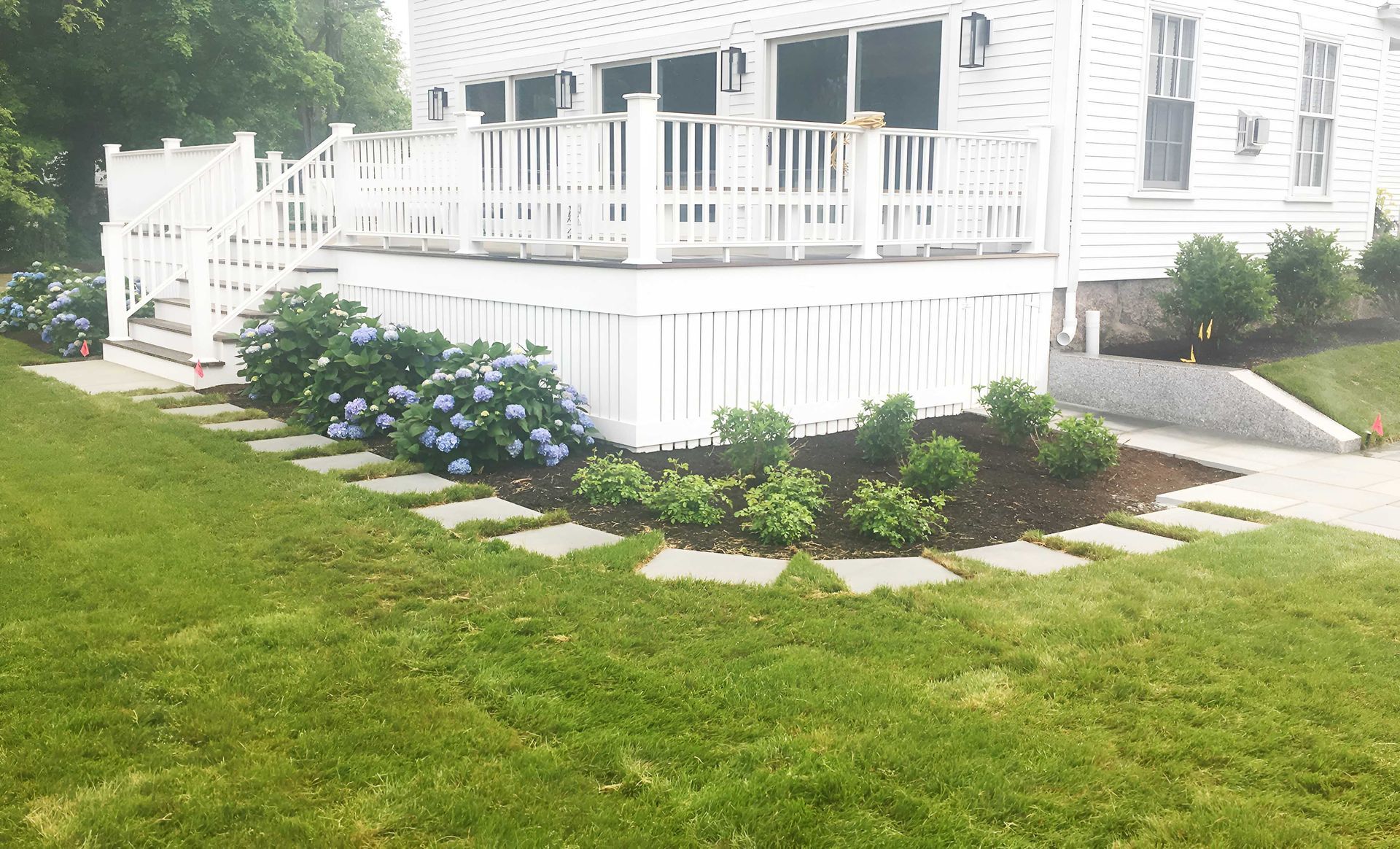 A white house with a raised wooden deck, featuring a landscaped garden with blue hydrangeas and stone path edging.