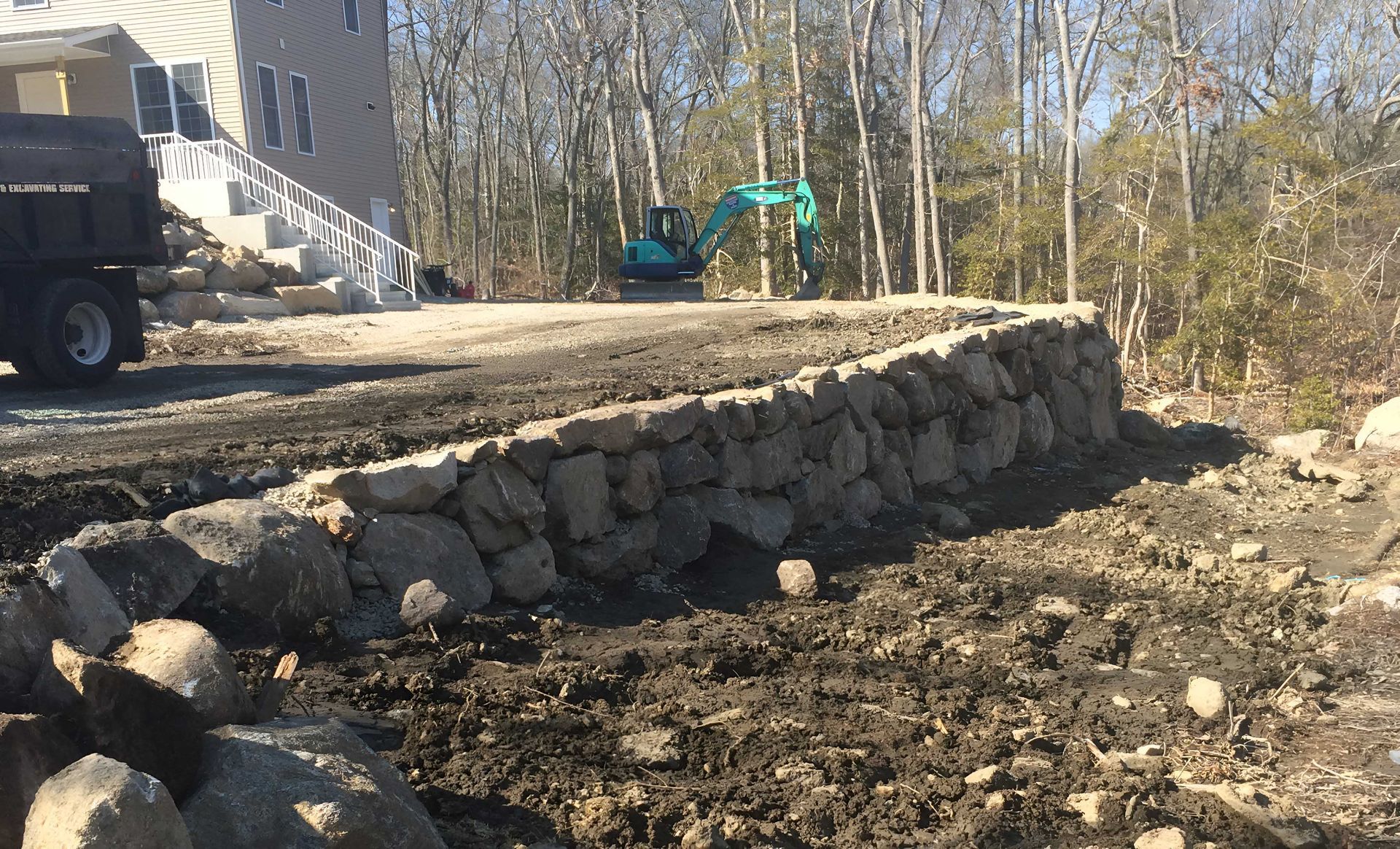 A house with a newly constructed stone retaining wall, with a teal excavator parked on the dirt lot behind it.