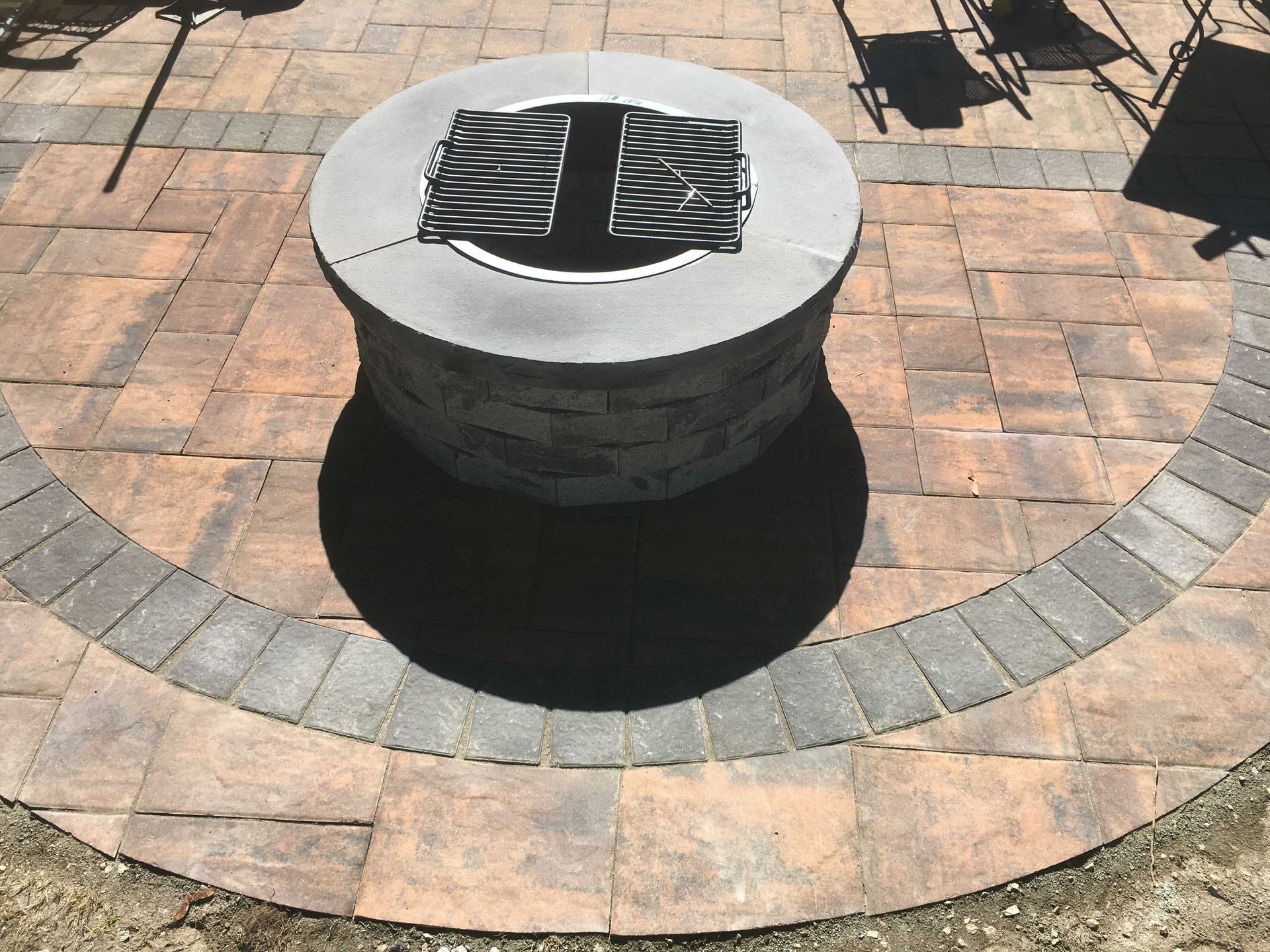 A round, grey stone fire pit sits in the center of a circular patterned patio made of tan and grey concrete pavers.