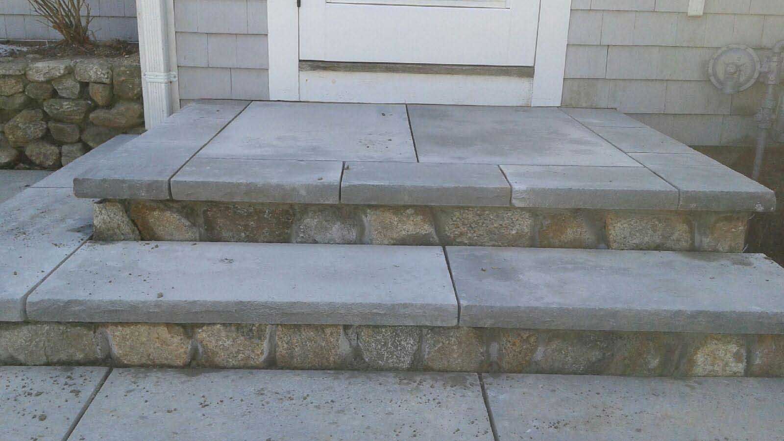 Two stone-faced porch steps leading to a white door, with flat grey stone treads and a textured rock base.