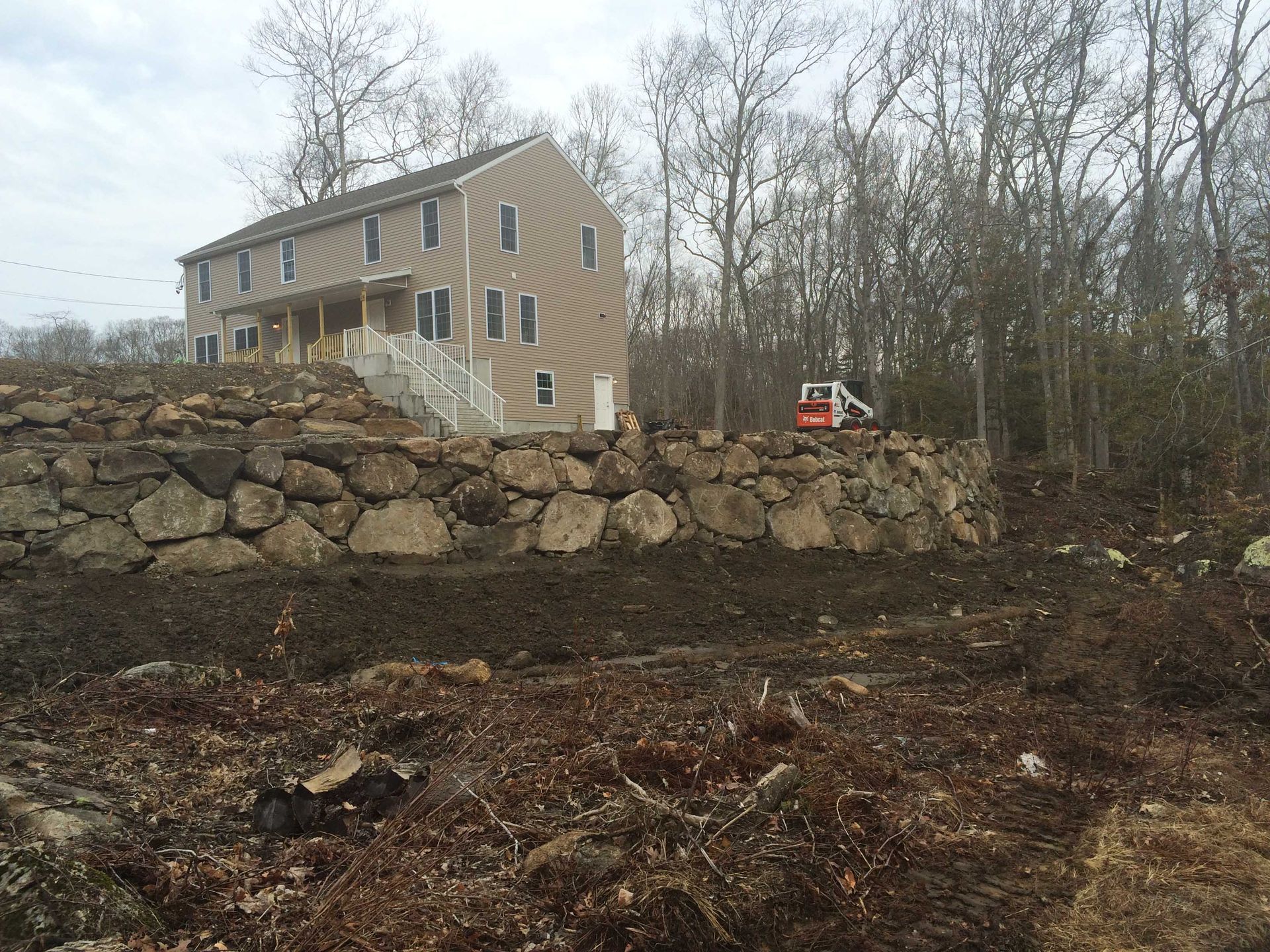 A large, beige, two-story house sits atop a stone retaining wall in a wooded area with a small excavator nearby.