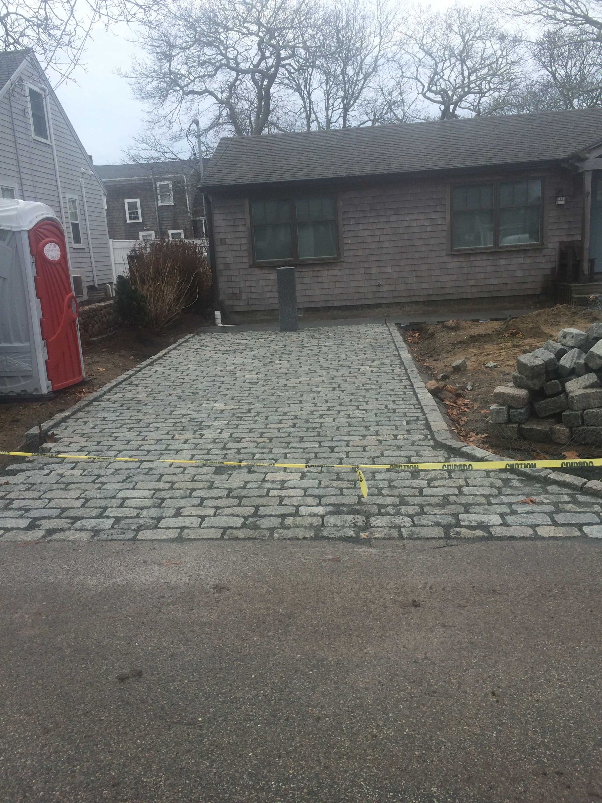 A newly laid cobblestone driveway leads to a brick house on a cloudy day, with a portable toilet visible to the side.